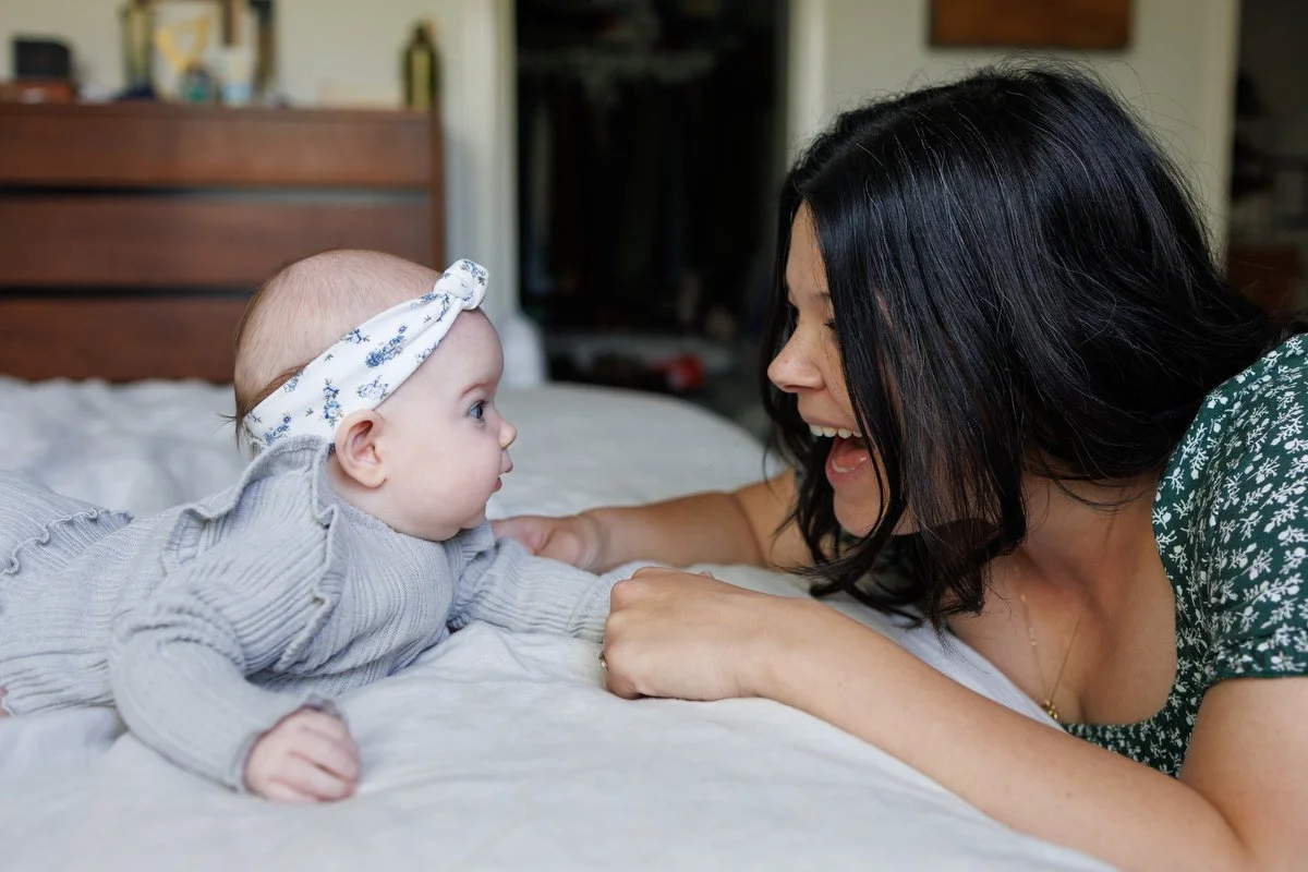 12-week-old baby in a grey onesie and headband plays with her mom, who is smiling and kneeling down on her level