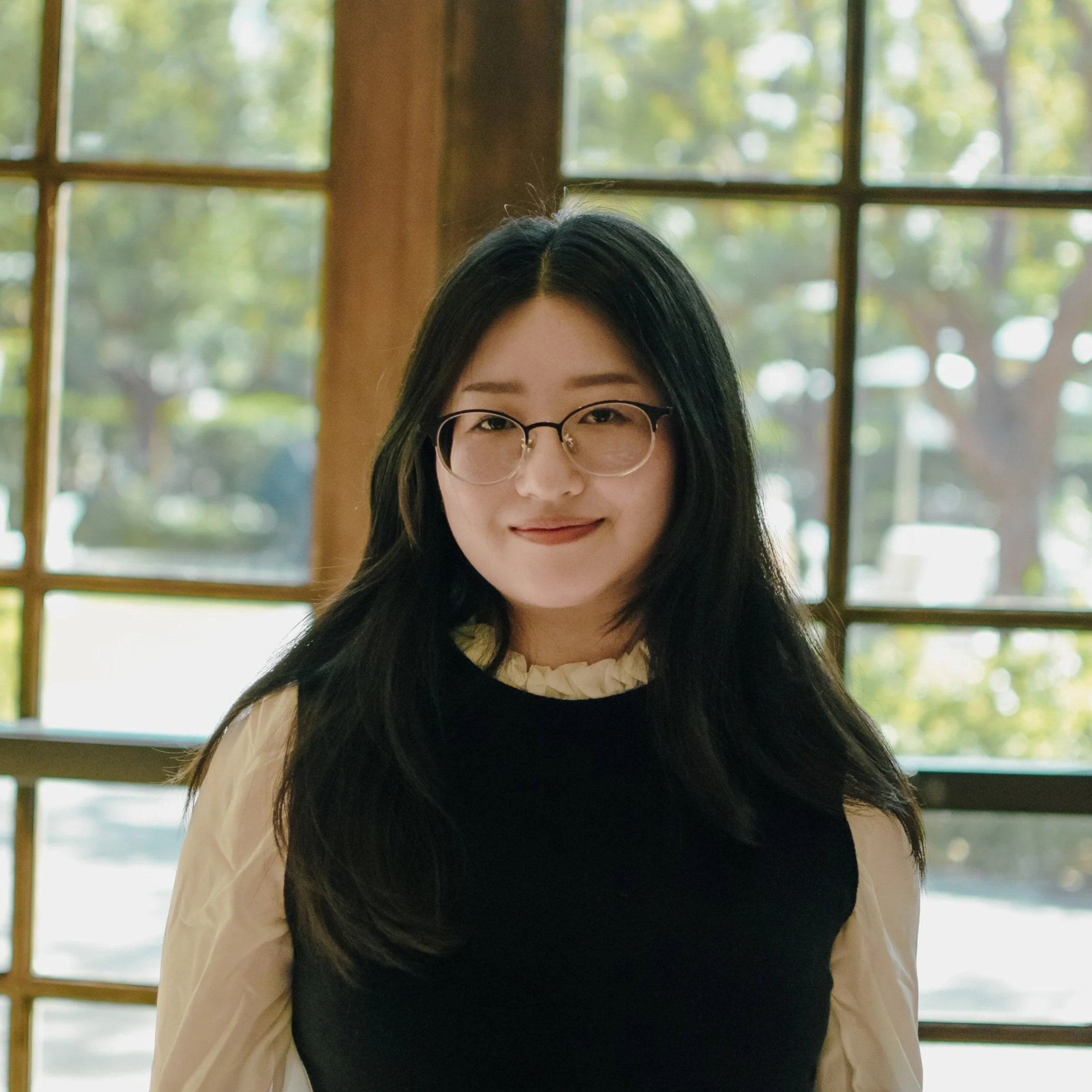 A professional woman with long dark hair, wearing a beige shirt and black blazer, standing indoors near a wooden wall and plants.