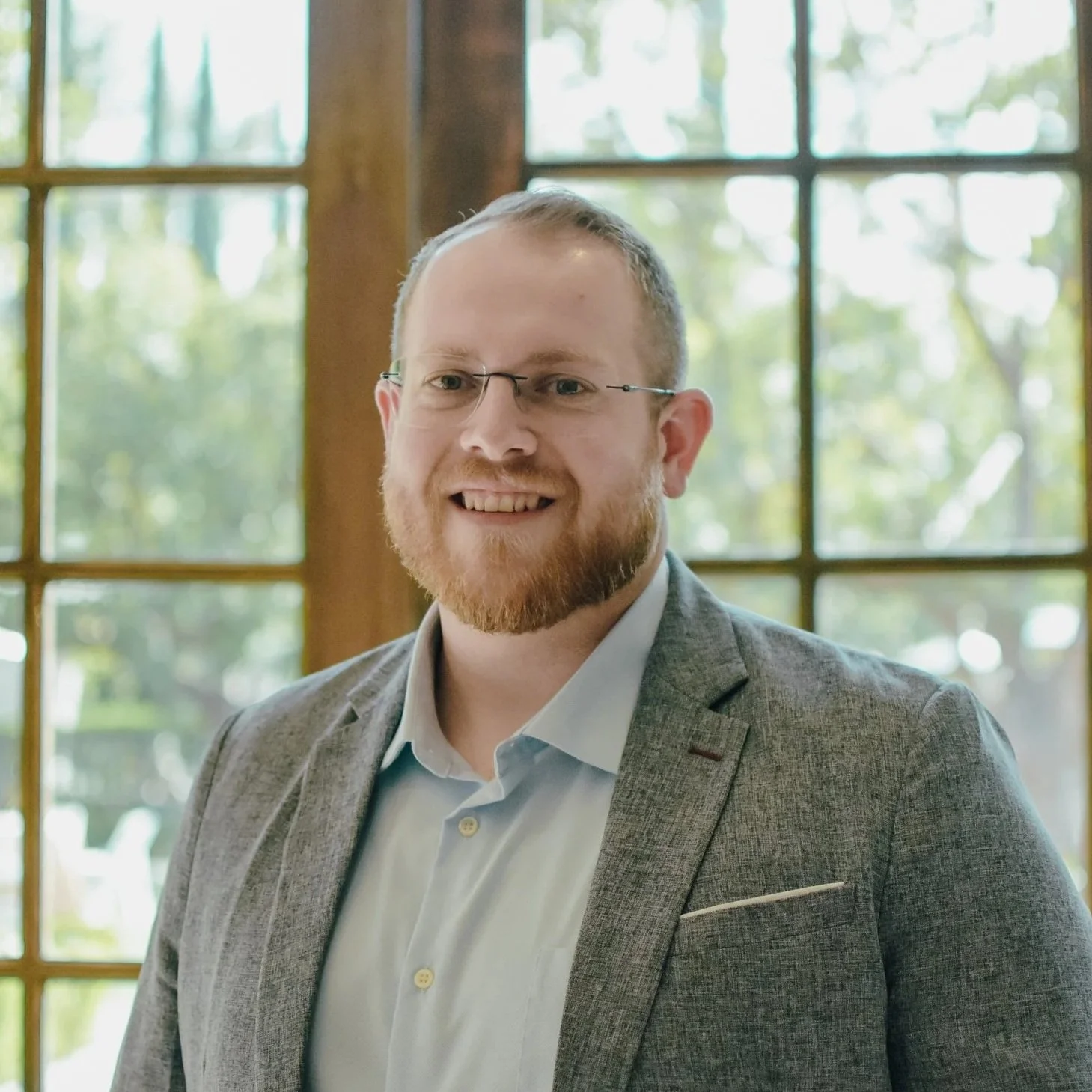 A smiling man with short hair and a beard wearing a light gray shirt, standing outdoors with a rocky, reddish-brown background.