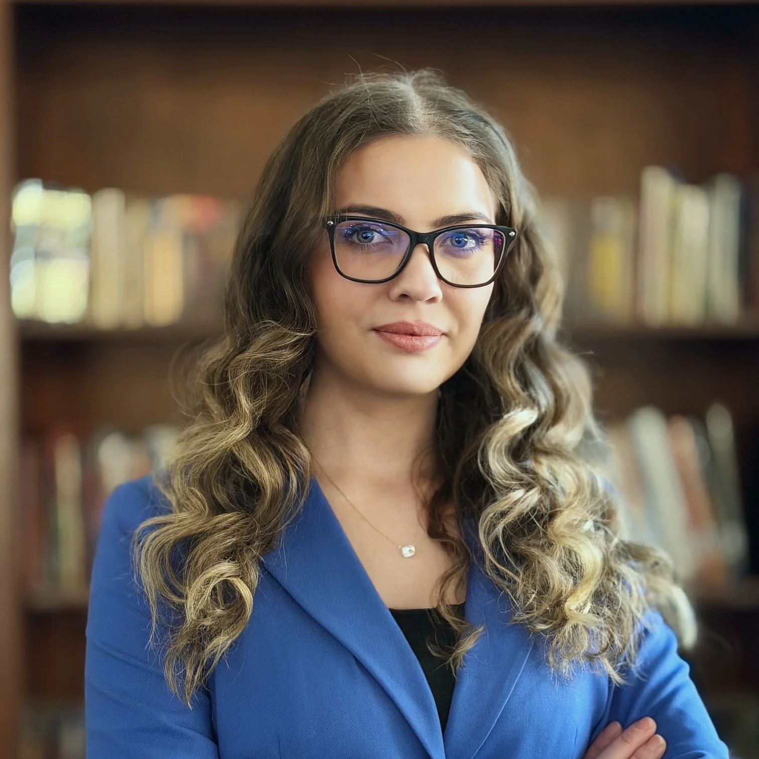 A young woman with long, wavy, blonde hair and blue eyes smiling while sitting in a room with warm lighting, a leather chair, and decorative wood branches in the background.