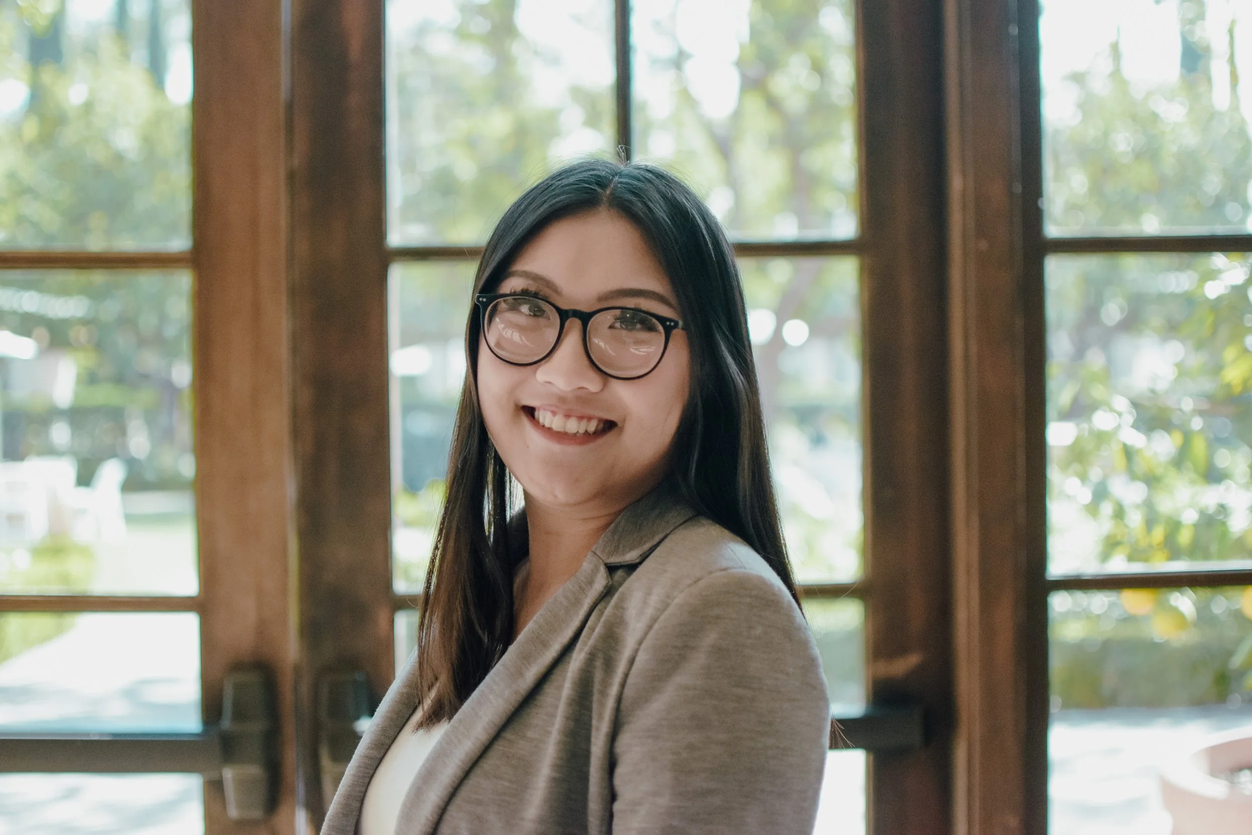 A young woman with long, straight dark hair smiling, wearing a black blazer, standing indoors against a background of wooden paneling and a white wall.