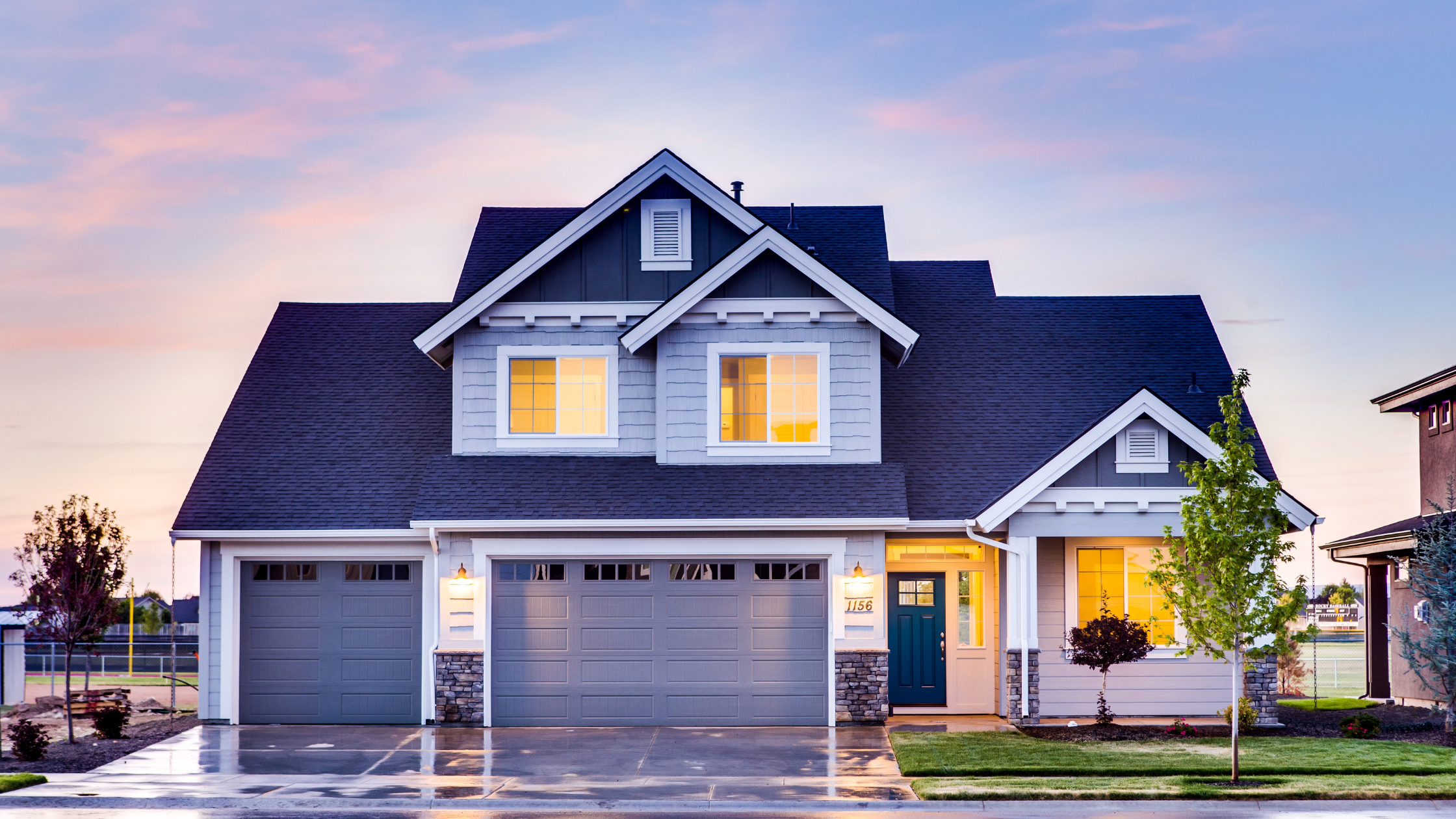 A modern two-story house with a dark gray roof, white exterior with gray and stone accents, illuminated by exterior lights, on a wet driveway at dusk.
