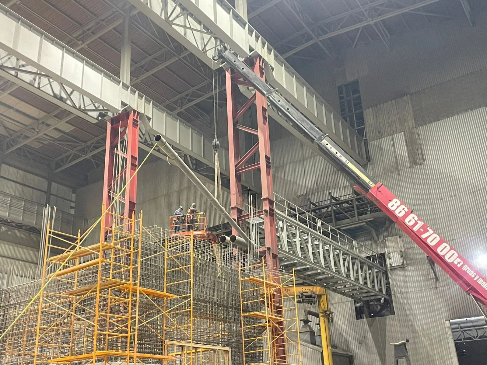 Trabajadores en un andamio en una construcción industrial con estructura de acero y grúas altas en un espacio cerrado.