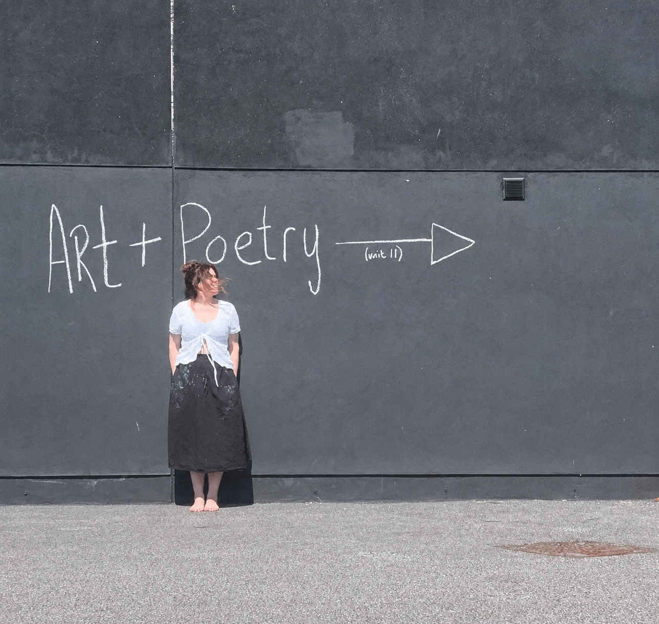 A woman with short hair stands barefoot against a dark wall with her hands in the pockets of her black skirt. The wall has a chalk drawing that reads 'Art + Poetry' with an arrow pointing to the right, labeled '(unit 11)'.