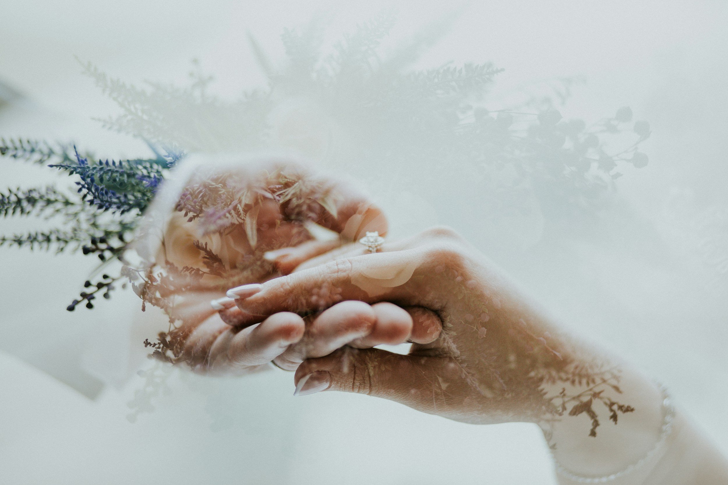 A close-up of two hands holding a small floral arrangement with greenery and flowers, captured through a window or glass surface with reflections of more greenery and flowers.