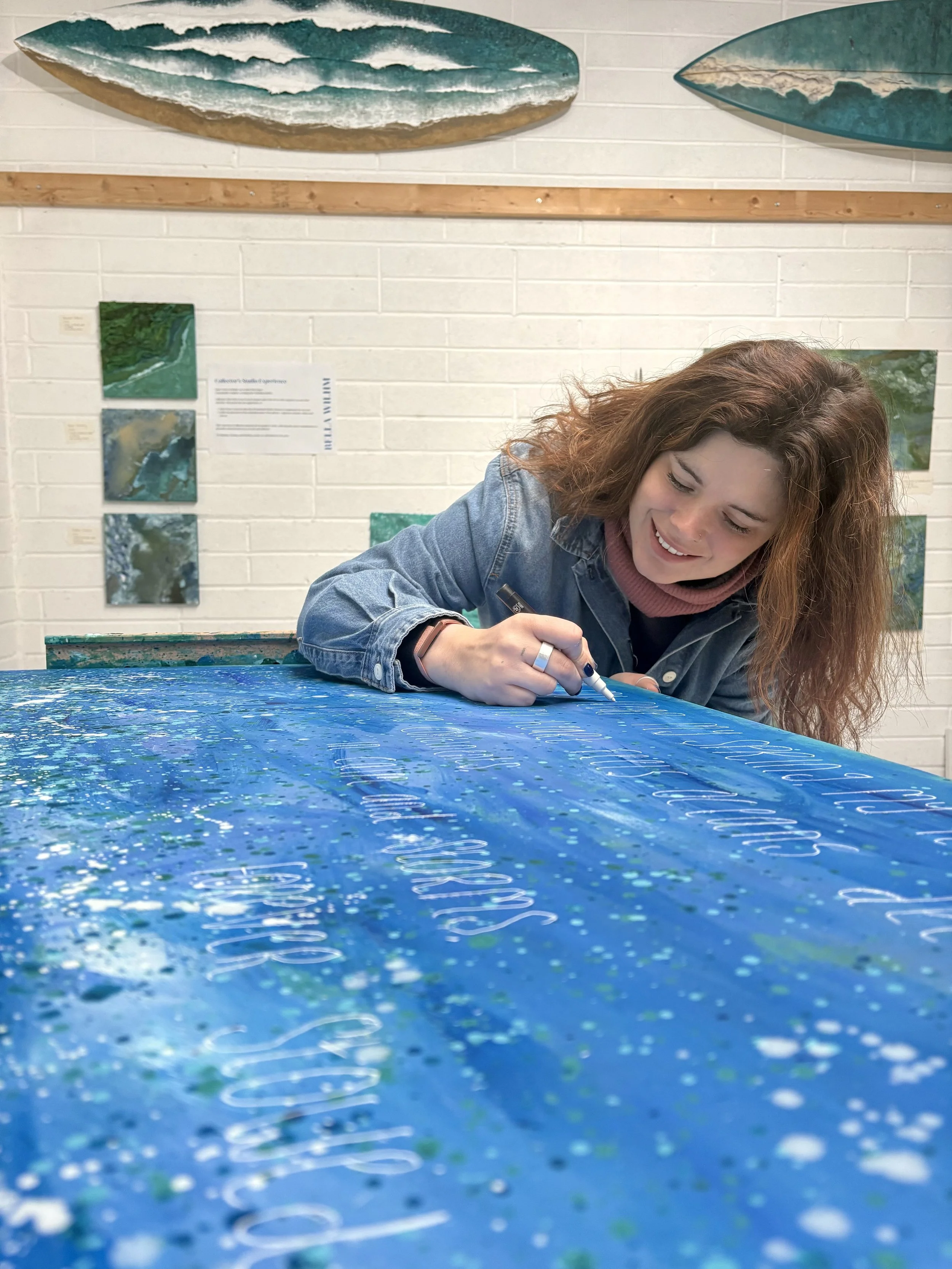 A woman with curly hair in a denim jacket writes on a blue, turquoise, and white ink-painted canvas with a marker. In the background, there are ocean-themed artwork and informational placards on the wall.