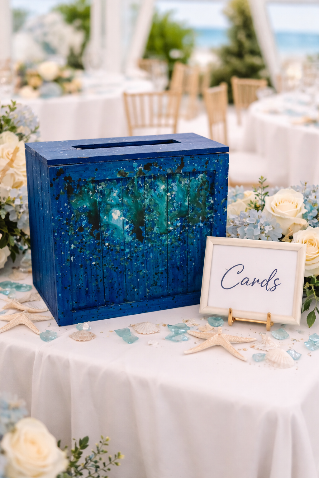 A decorative blue box with an artistic splatter design, surrounded by flowers, seashells, starfish, and a sign labeled 'Cards,' at a wedding or event table.