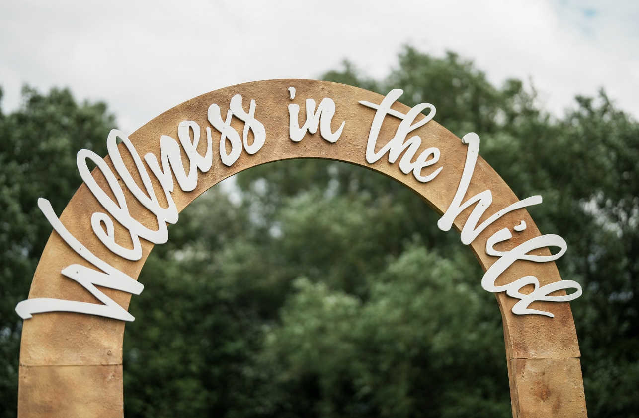 Wooden arch with white cursive sign reading 'Welcoming in the Wild' against a background of trees and cloudy sky.