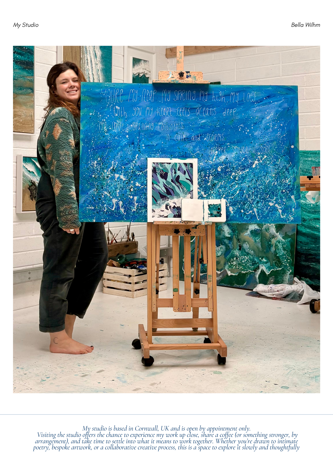 An artist standing barefoot in an art studio, holding a large blue abstract painting with handwritten text, surrounded by other abstract artworks and art supplies.