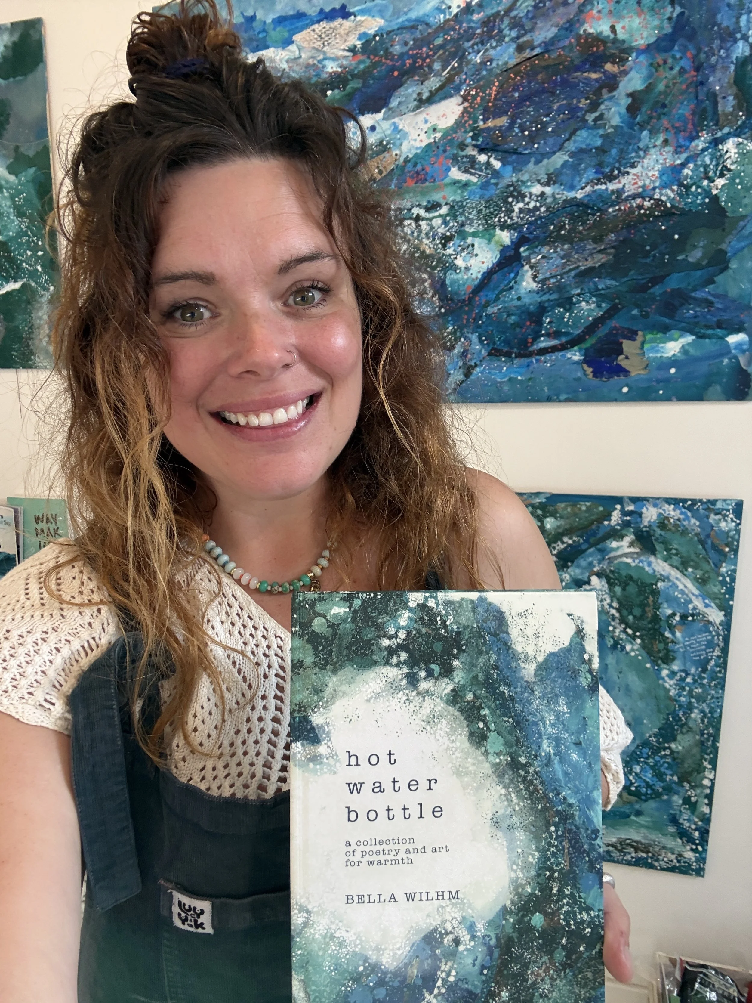 A woman with curly brown hair smiling, wearing a crochet top and beaded necklace, holding a book titled 'hot water bottle' with a background of abstract blue and green paintings.