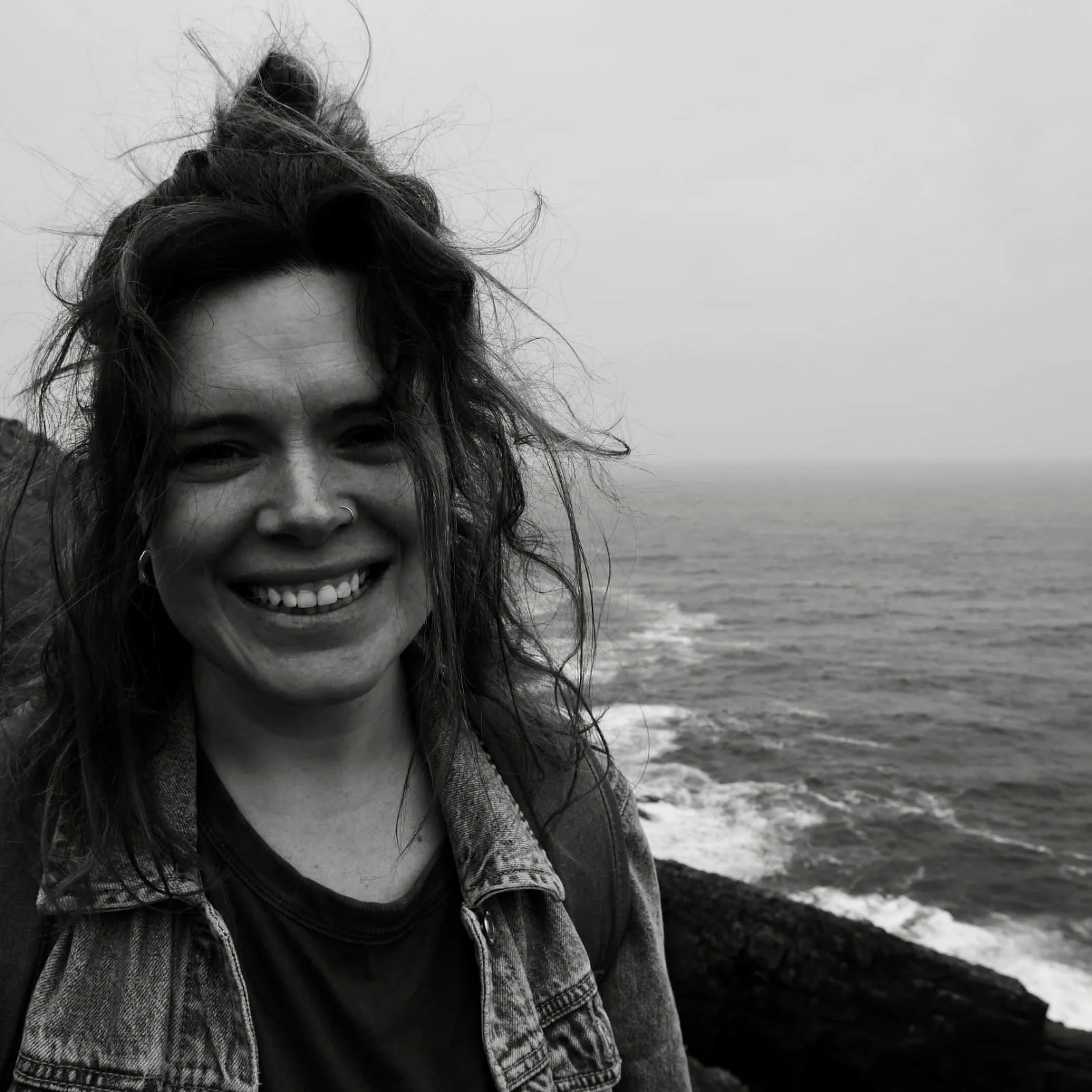 Black and white photo of a smiling woman with curly hair and a nose ring, standing near the ocean with rocky shoreline in the background.