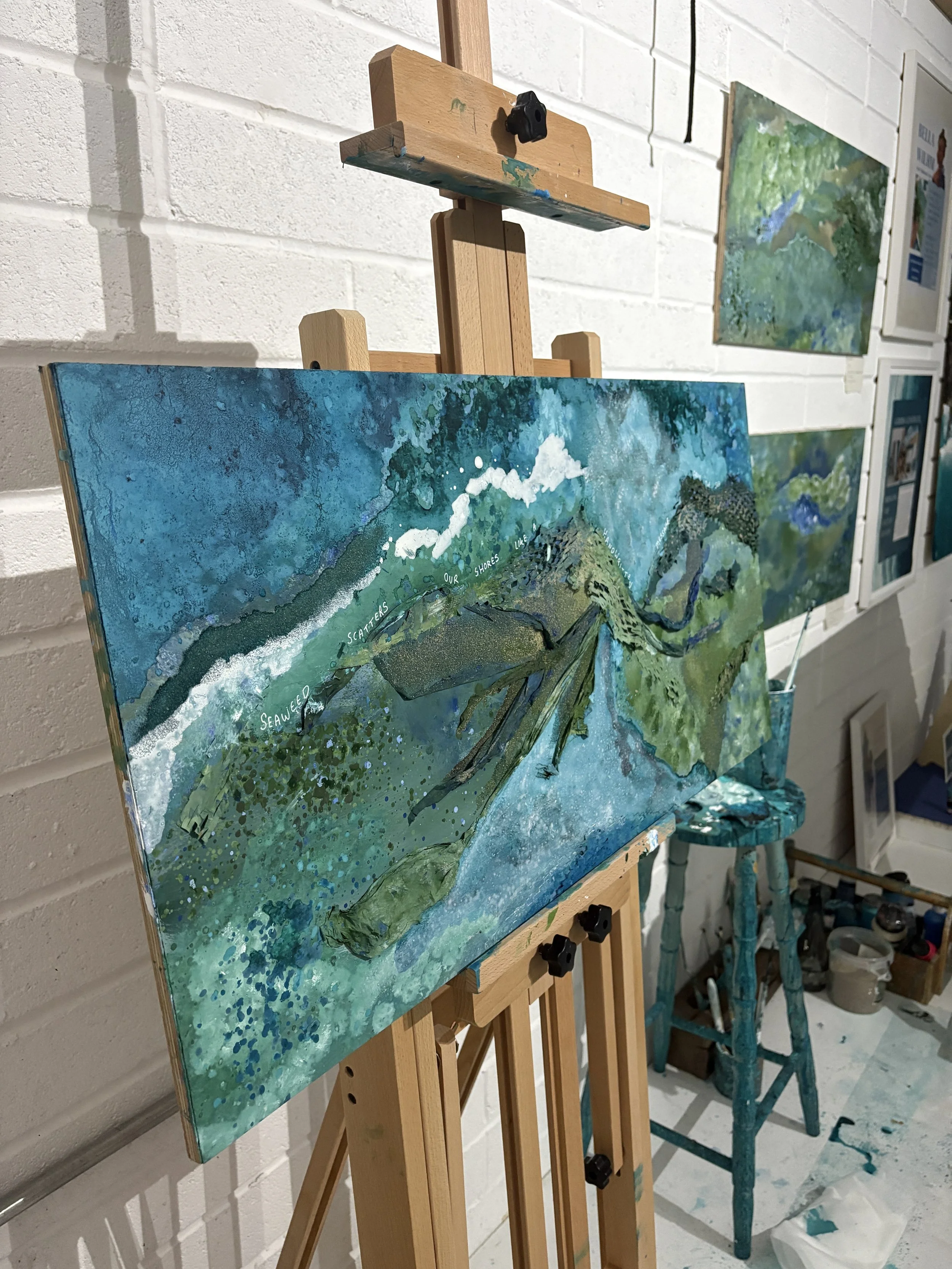A colorful abstract painting of a ocean landscape on an easel in an art studio. The painting contains blue, green, and white colours with the words 'Seaweed Scatters Our Shores" written across it.