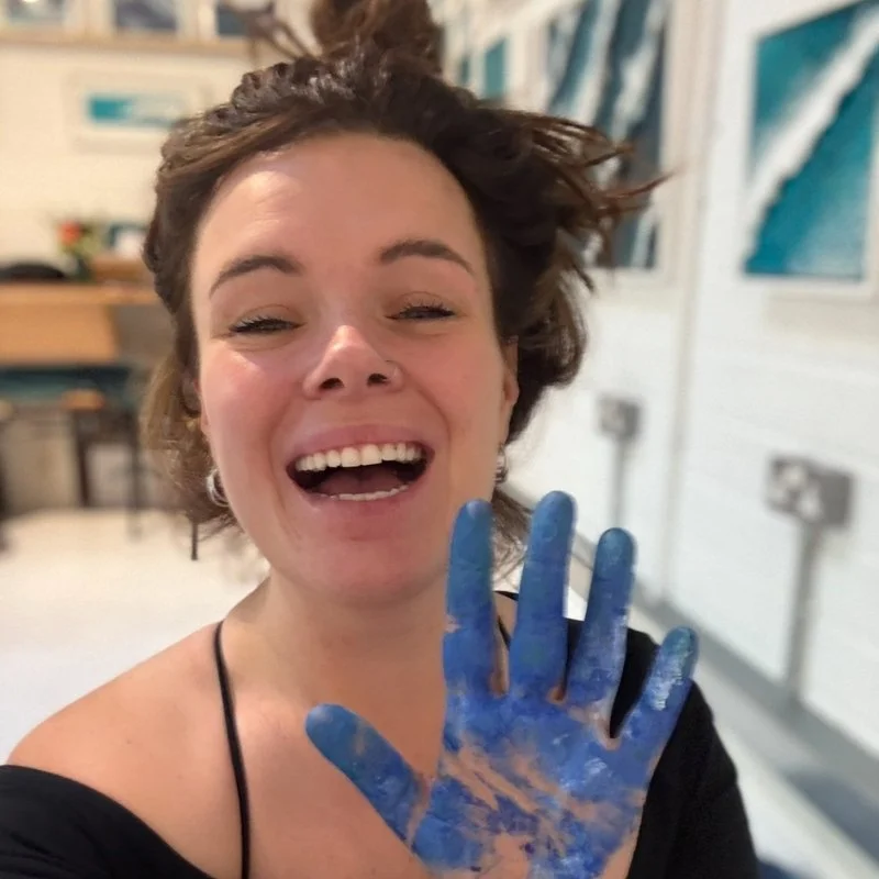 A woman with her brown hair in a bun, smiling and holding her hand up, covered in blue paint, inside an art studio in Cornwall