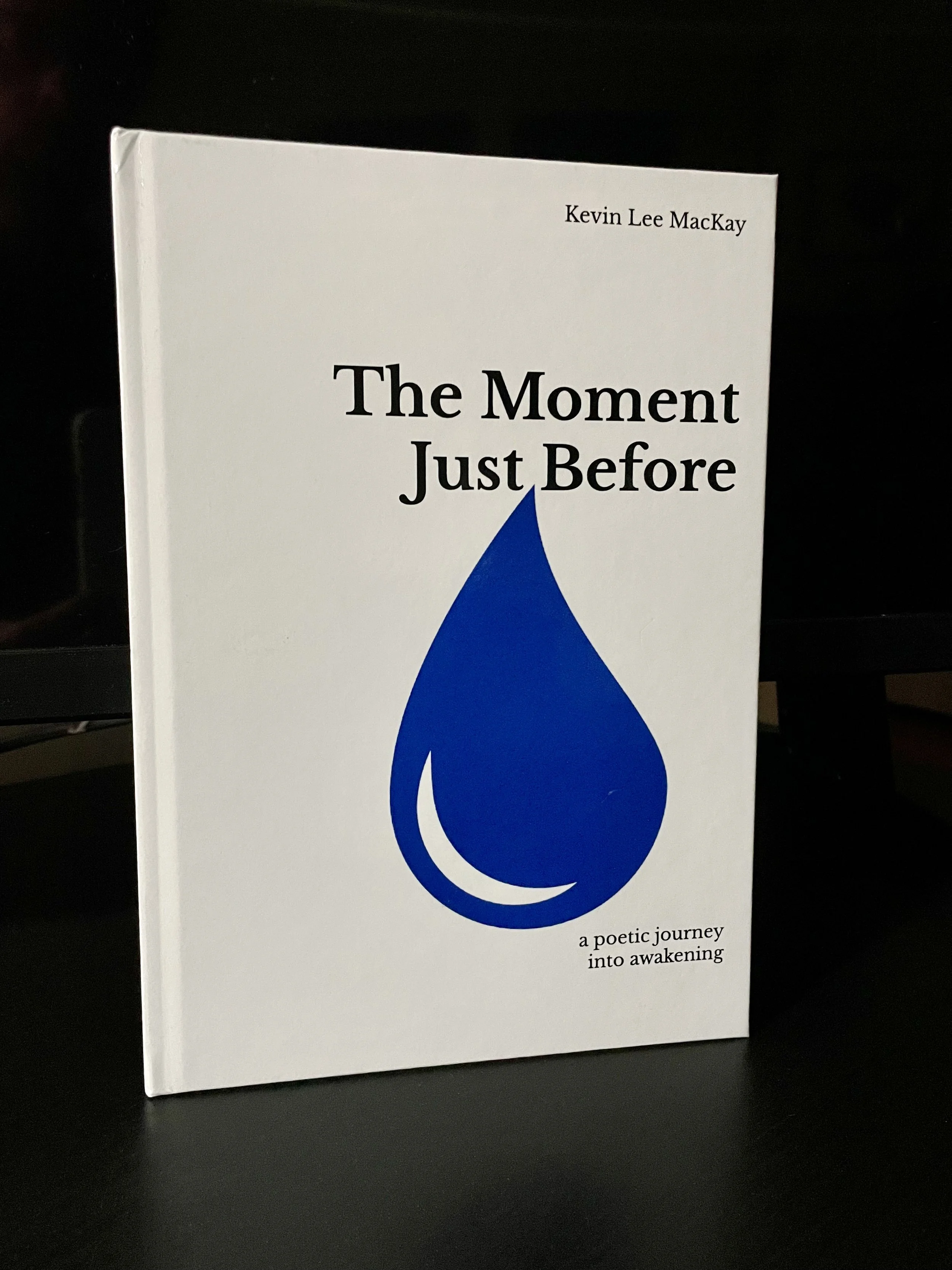 A white book cover titled 'The Moment Just Before' by Kevin Lee MacKay, with a large blue teardrop shape and the subtitle 'a poetic journey into awakening'.
