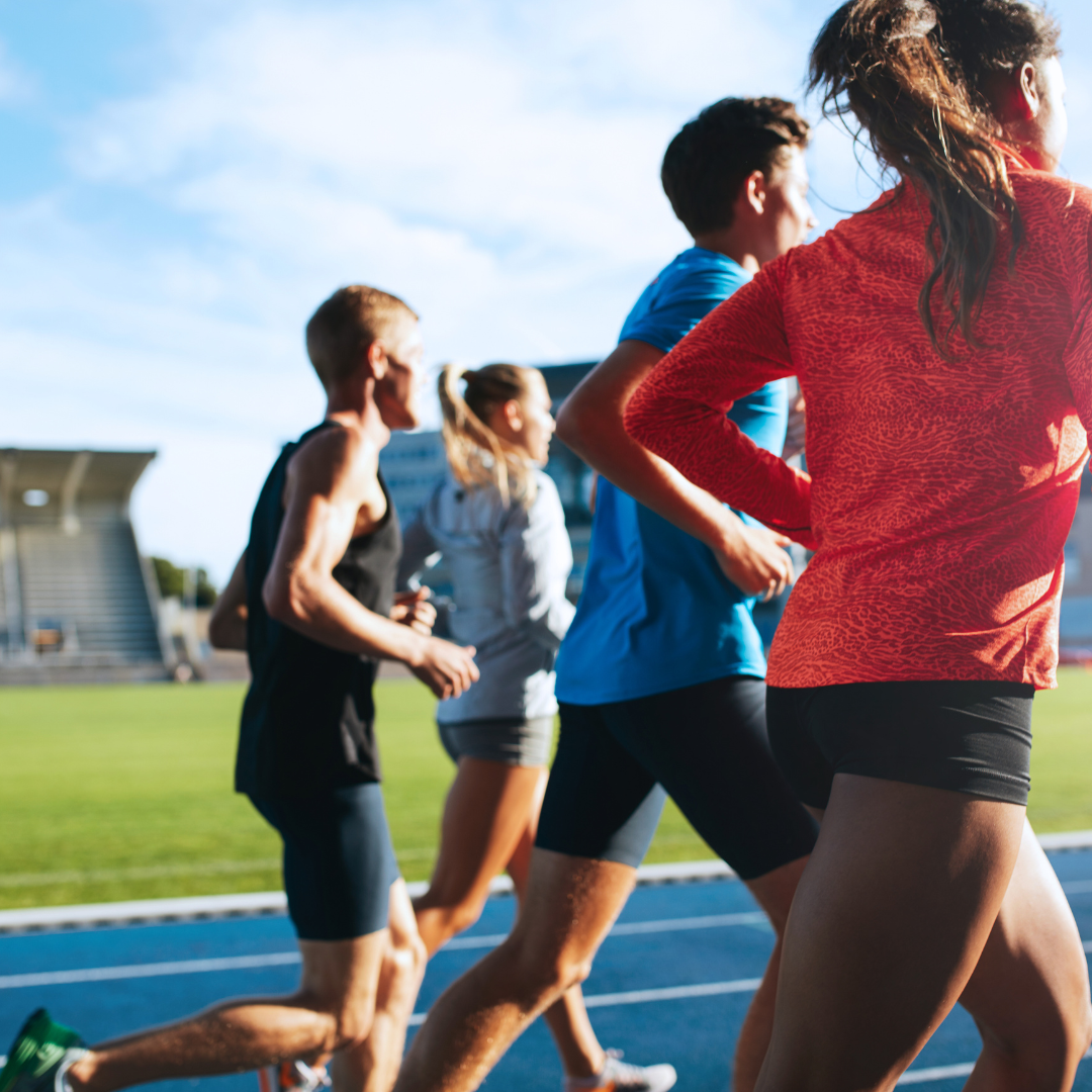Group of athletes running on a track outdoors, with stadium seats and clear sky in the background.