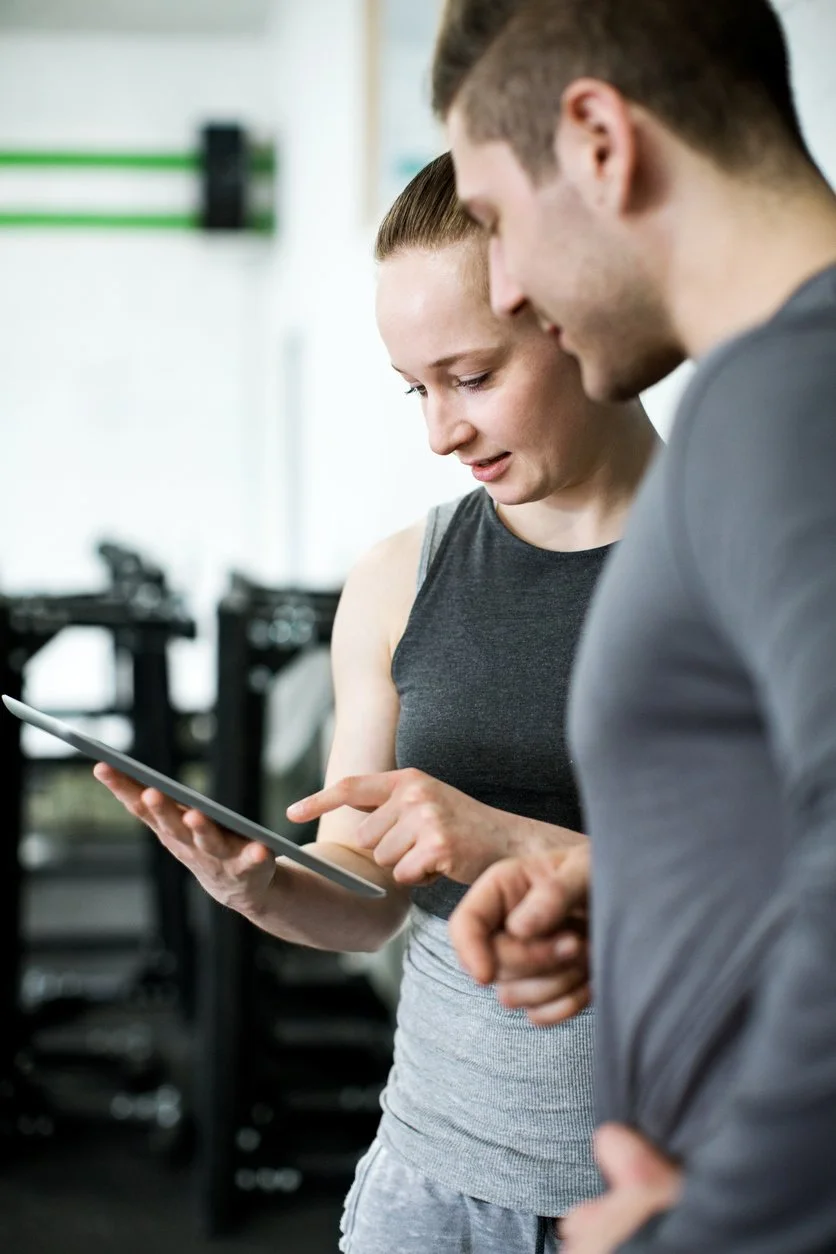 Two people look at a tablet together in a gym setting, one of them pointing at the screen. Exercise equipment is visible in the background.