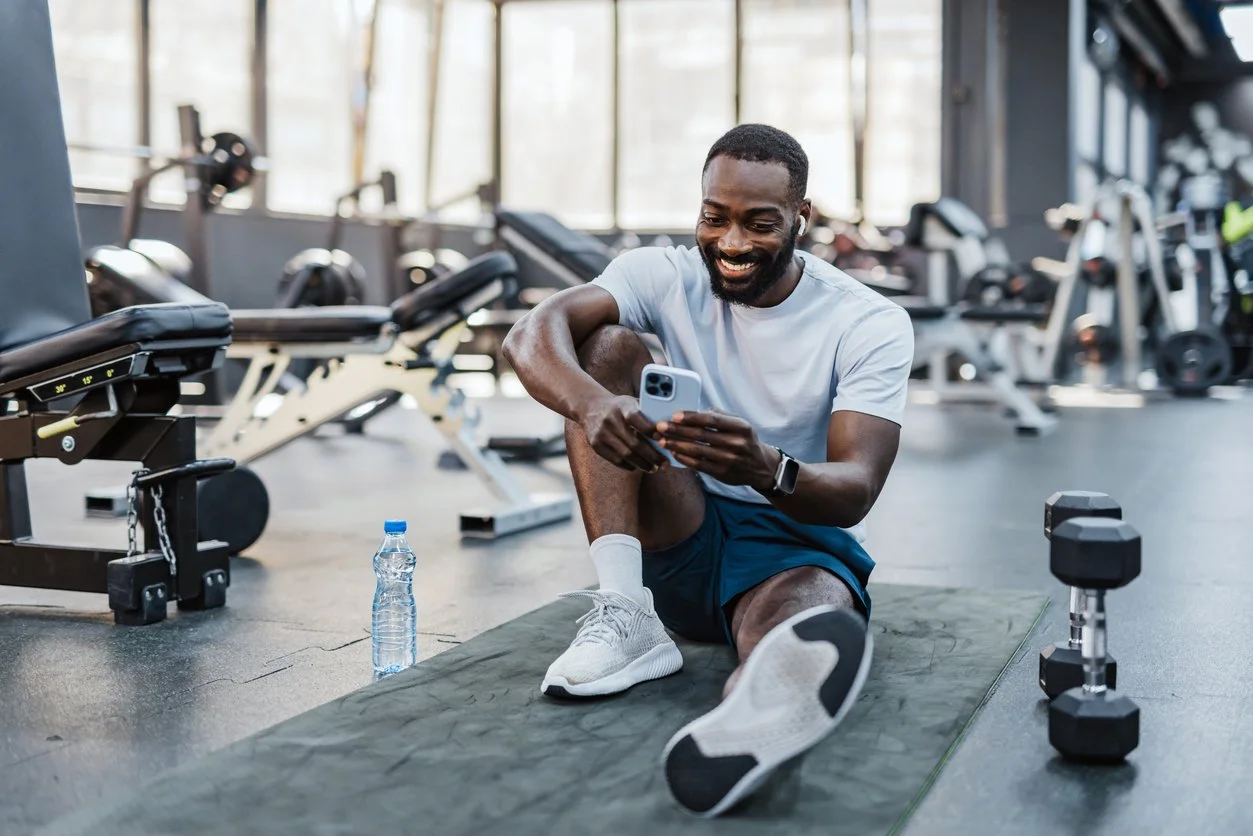 Man in workout attire sitting on a mat at a gym, looking at his phone, with a water bottle and dumbbell nearby.