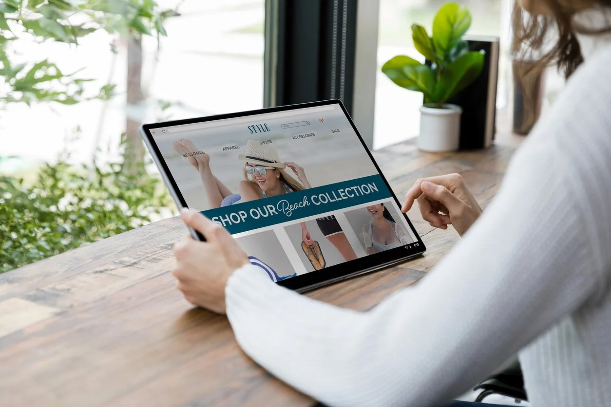 Person browsing an online beach clothing collection on a tablet at a wooden table with a plant and window in the background.