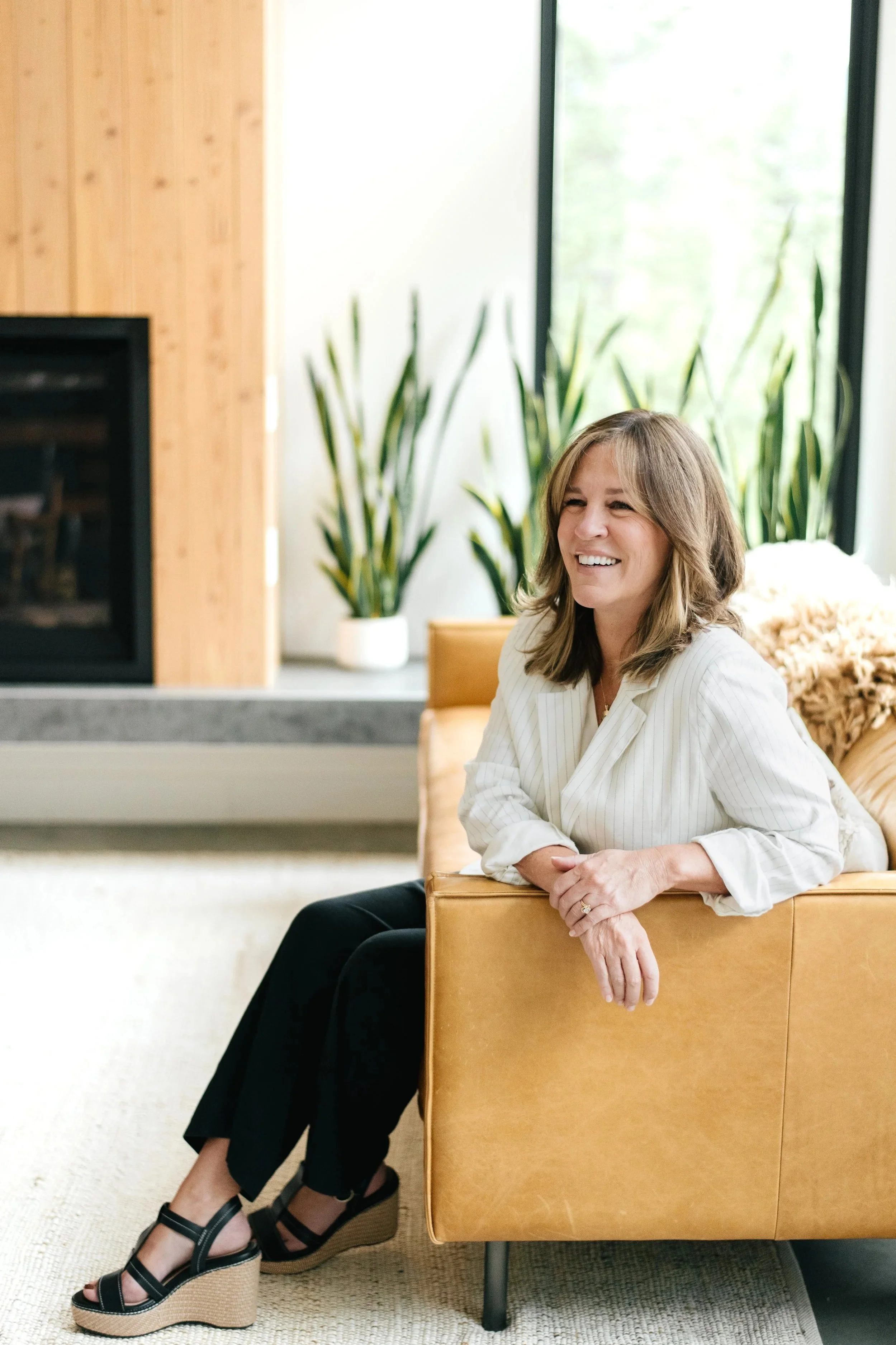 A woman with shoulder-length hair smiling and sitting relaxed on a beige leather couch in a bright, modern living room with large windows, green plants, and a fireplace.
