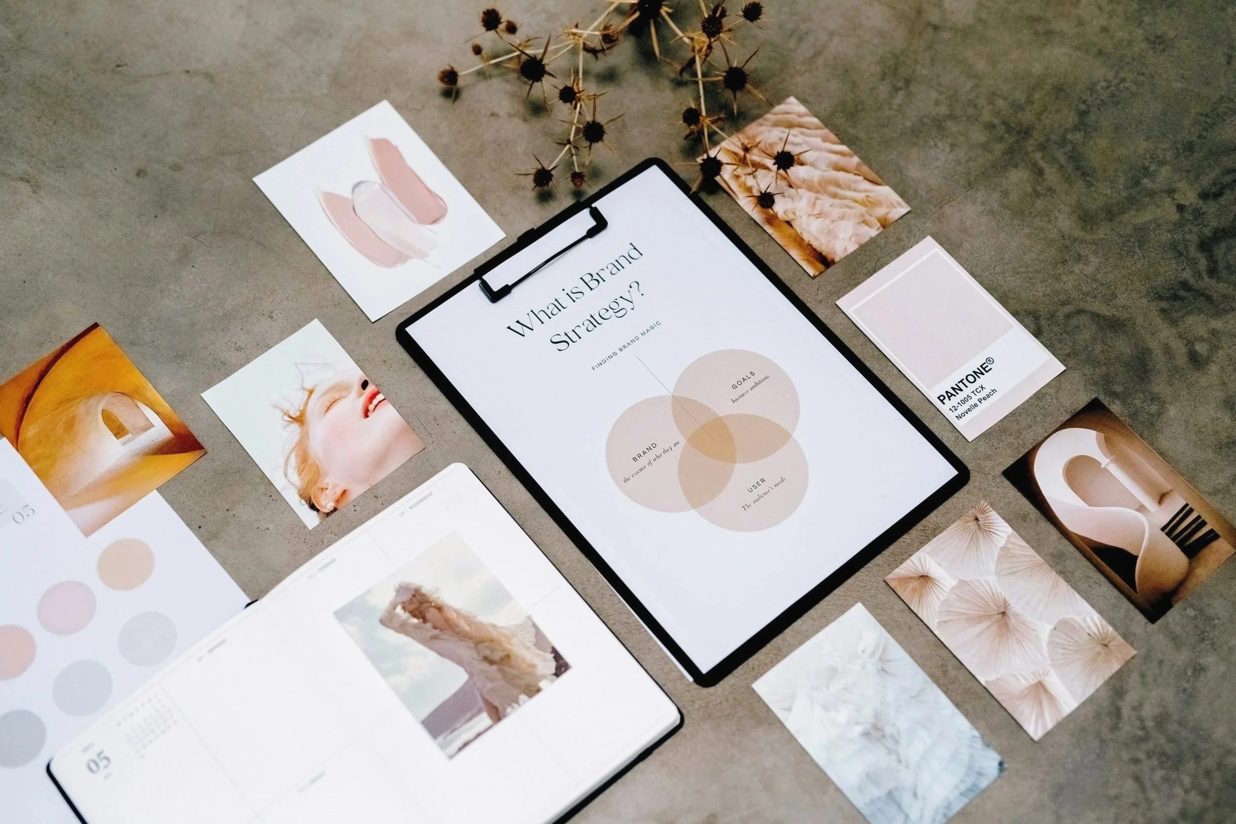 A flat lay of branding materials on a concrete surface, including a tablet displaying "What is Brand Strategy?" with Venn diagram, several watercolor-style images and photographs, color palette samples, and dried flowers.