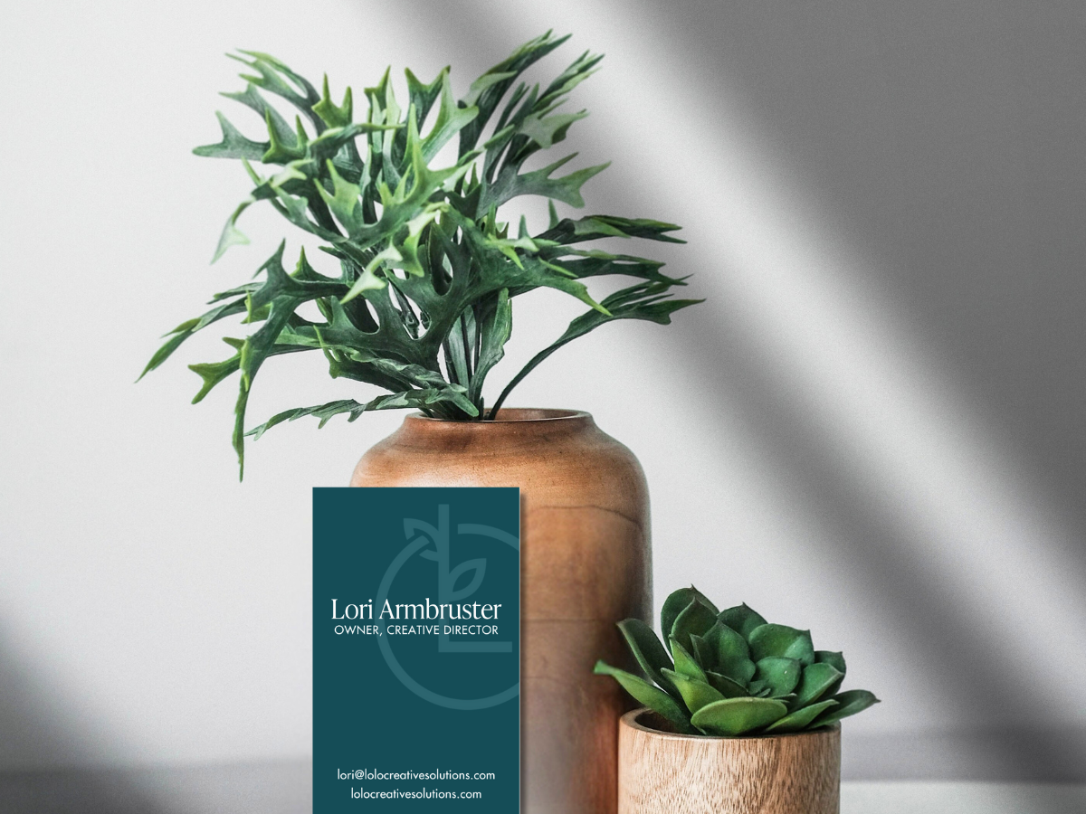 Two potted succulent plants, one in a taller brown pot with green leafy foliage, and the other in a smaller light-colored wooden pot, with a business card for Lori Ambruster, owner and creative director, leaning against the taller pot.