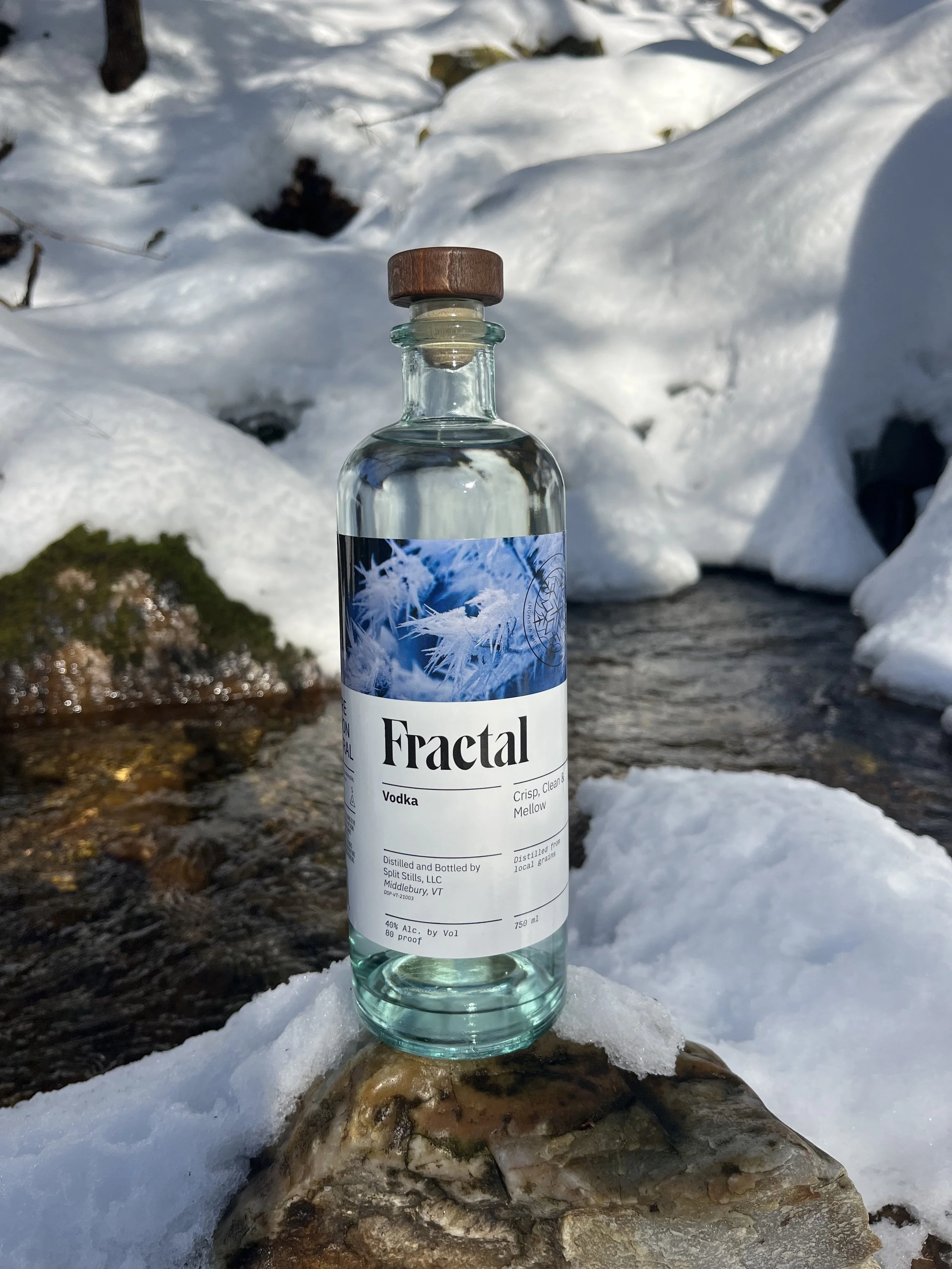 A clear bottle of Fractal vodka with a wooden cap, placed on a rock in a snowy outdoor setting, with snow-covered rocks and water in the background.