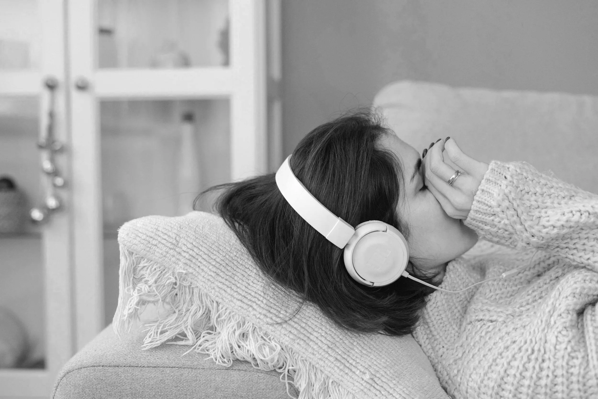 A woman with dark hair lying on a couch, wearing large headphones, with her eyes closed and hands resting on her forehead, suggesting relaxation or listening to music in a cozy environment.