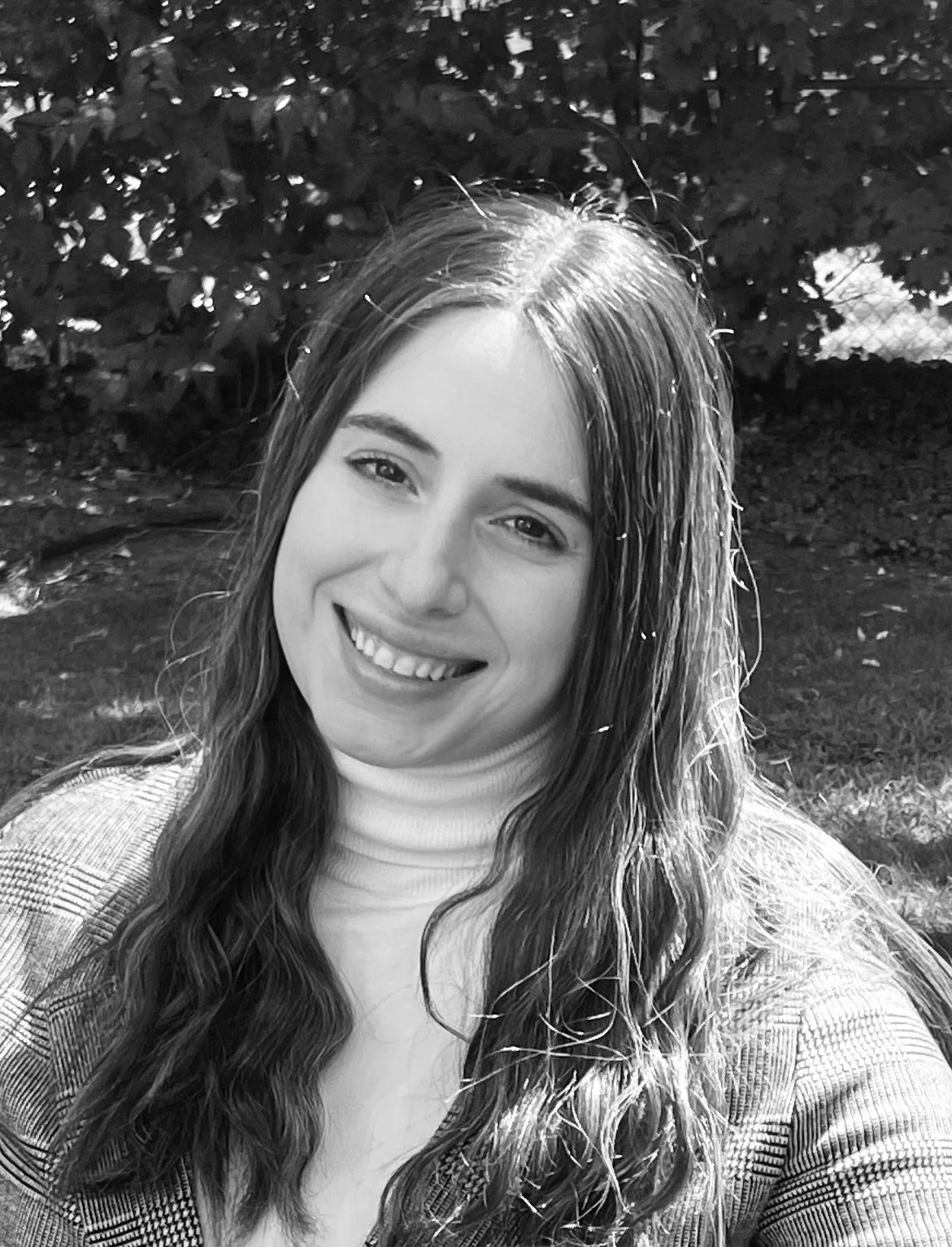 A young woman smiling outdoors with trees in the background.