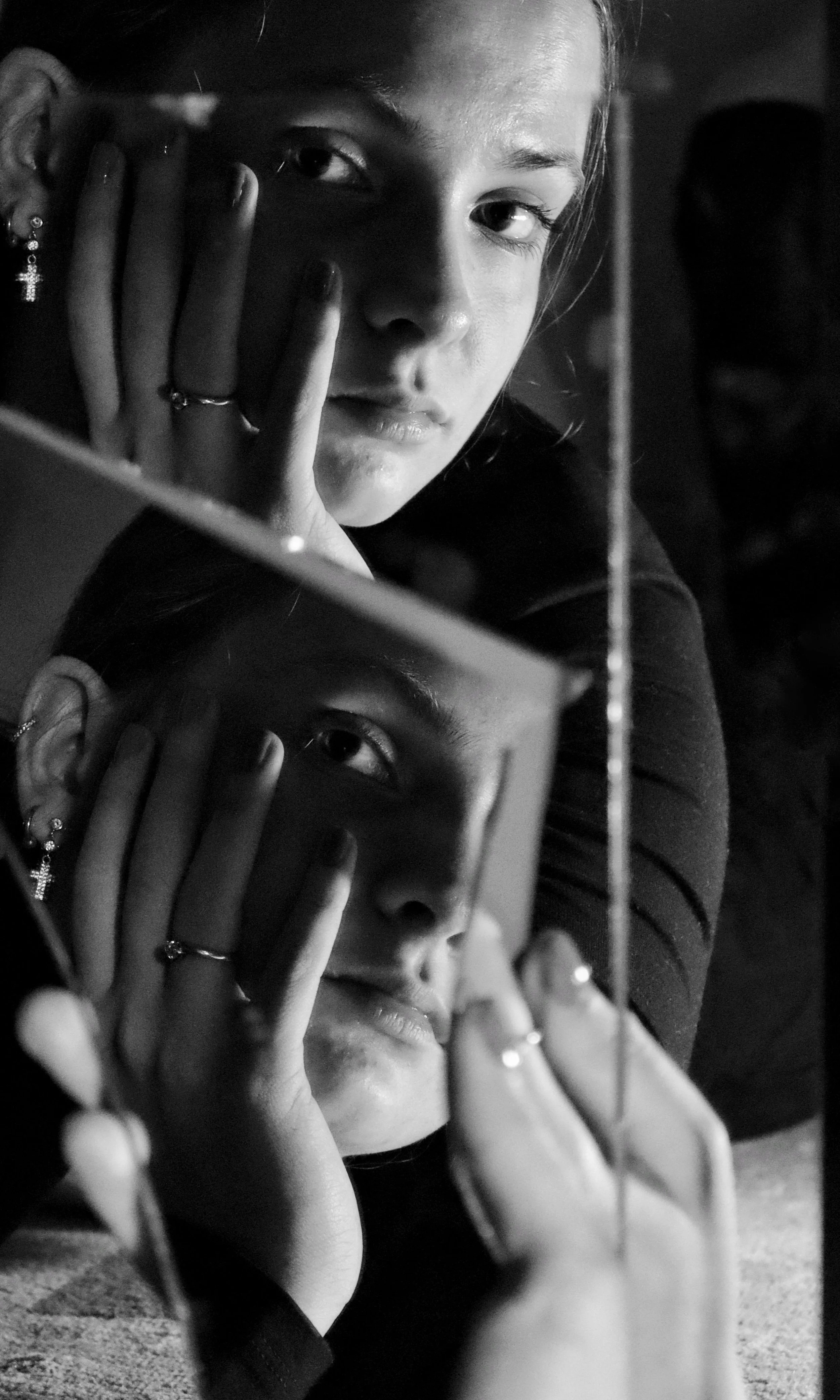 Close-up black and white photograph of a young woman with her face reflected in a mirror. She has earrings, rings, and is resting her hand on her face, with her eyes looking directly at the camera.