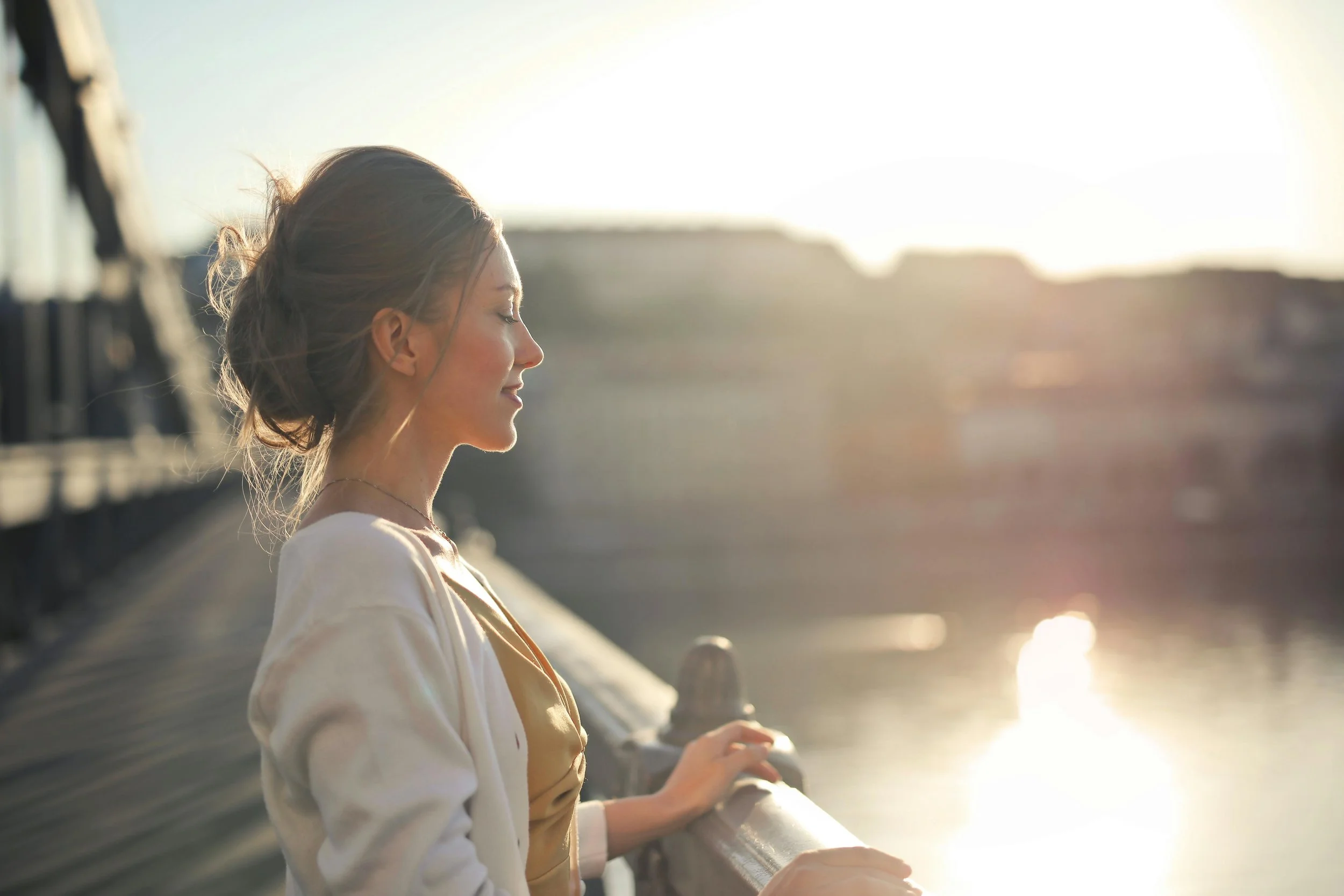 A young woman with closed eyes and a gentle smile, sitting on a bridge with a railing, enjoying the warm sunlight during sunset or sunrise, with blurred landscape in the background.