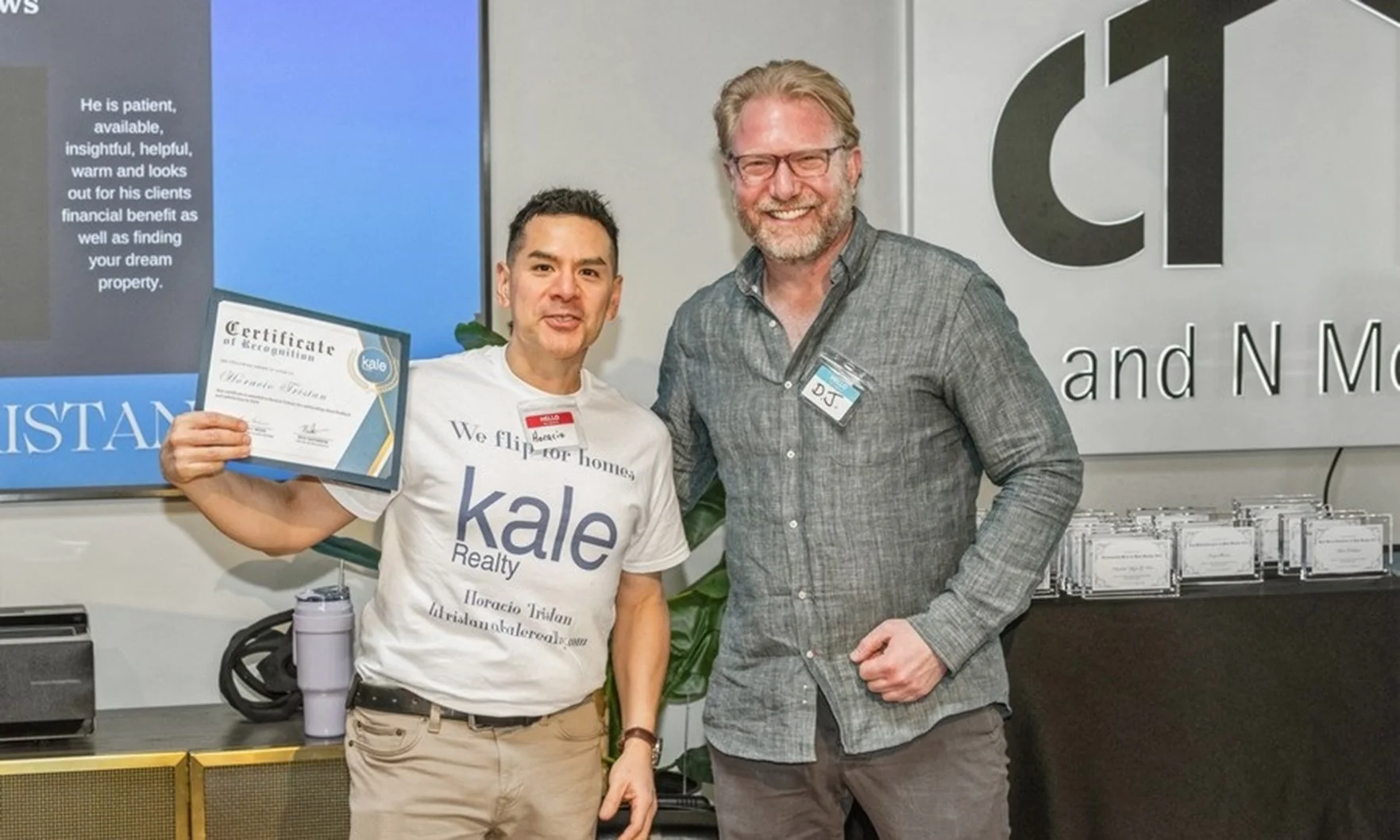 Two men smiling, one holding a certificate, at an indoor event with a display board and table.