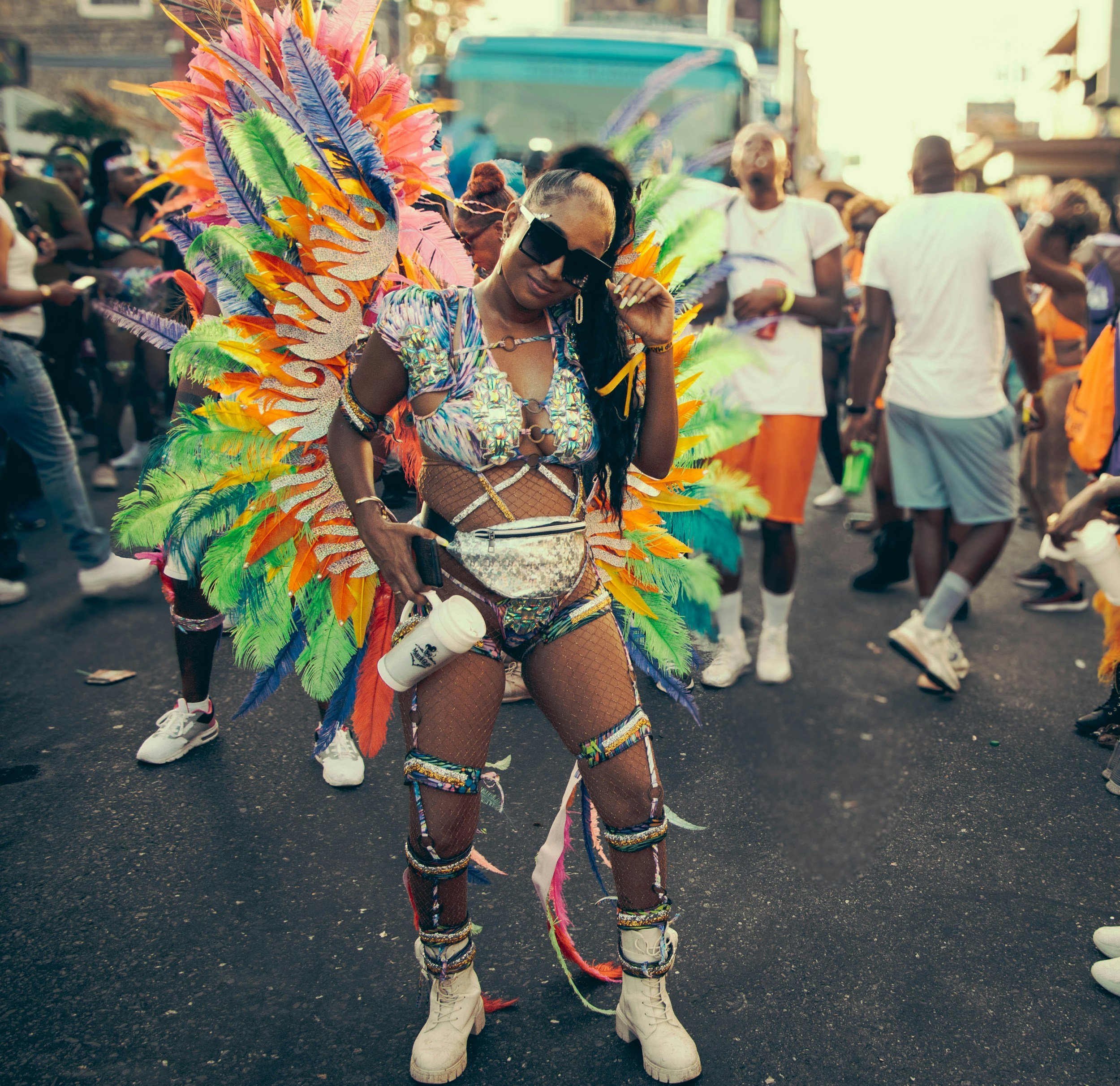 A woman dancing at a street carnival wearing a colorful feathered costume and sunglasses, surrounded by other festively dressed people.
