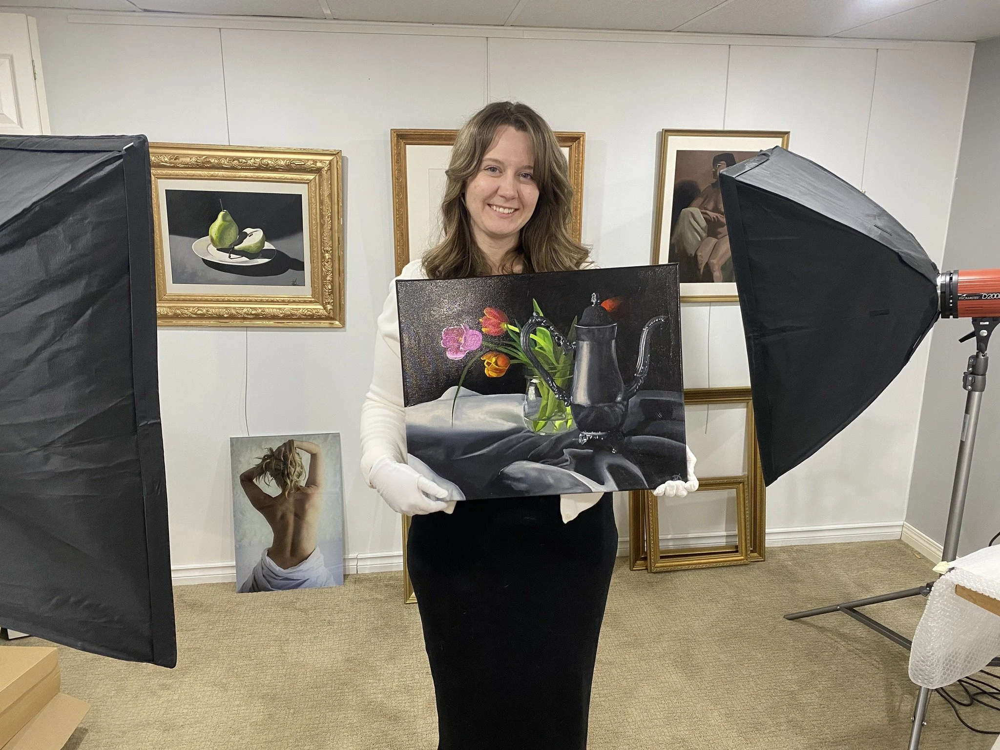 A woman in a white blazer and black skirt holding a still life painting of flowers and a vintage coffee pot in an art gallery or studio, with various framed artworks on the wall and professional photography lighting equipment in the scene.