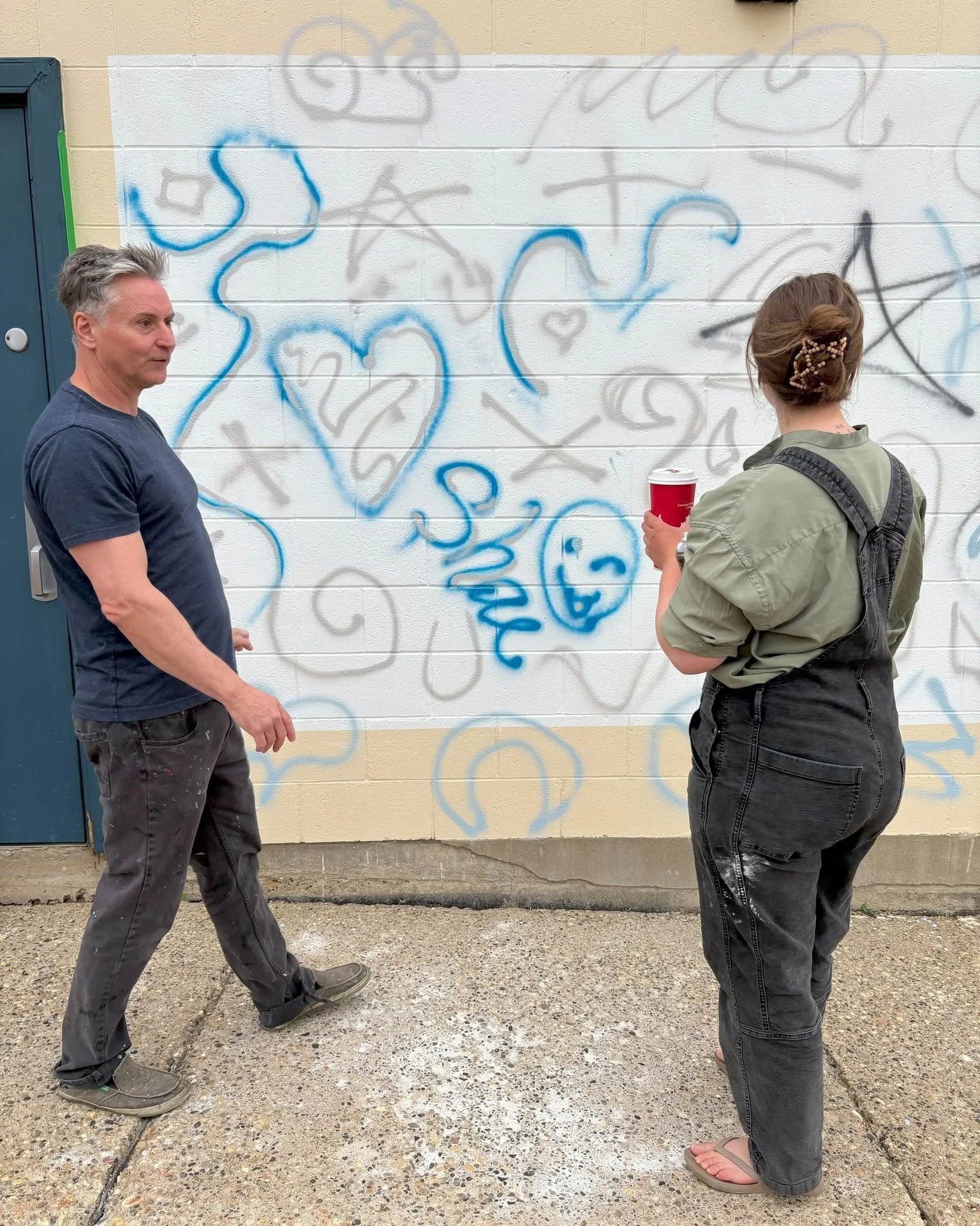 Two people standing in front of a wall with graffiti, one man and one woman, holding a red cup, engaged in conversation.
