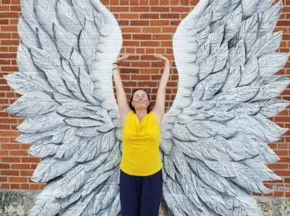 Lydia smiling with eyes closed, hands stretched to the sky with angel wings on the wall behind her