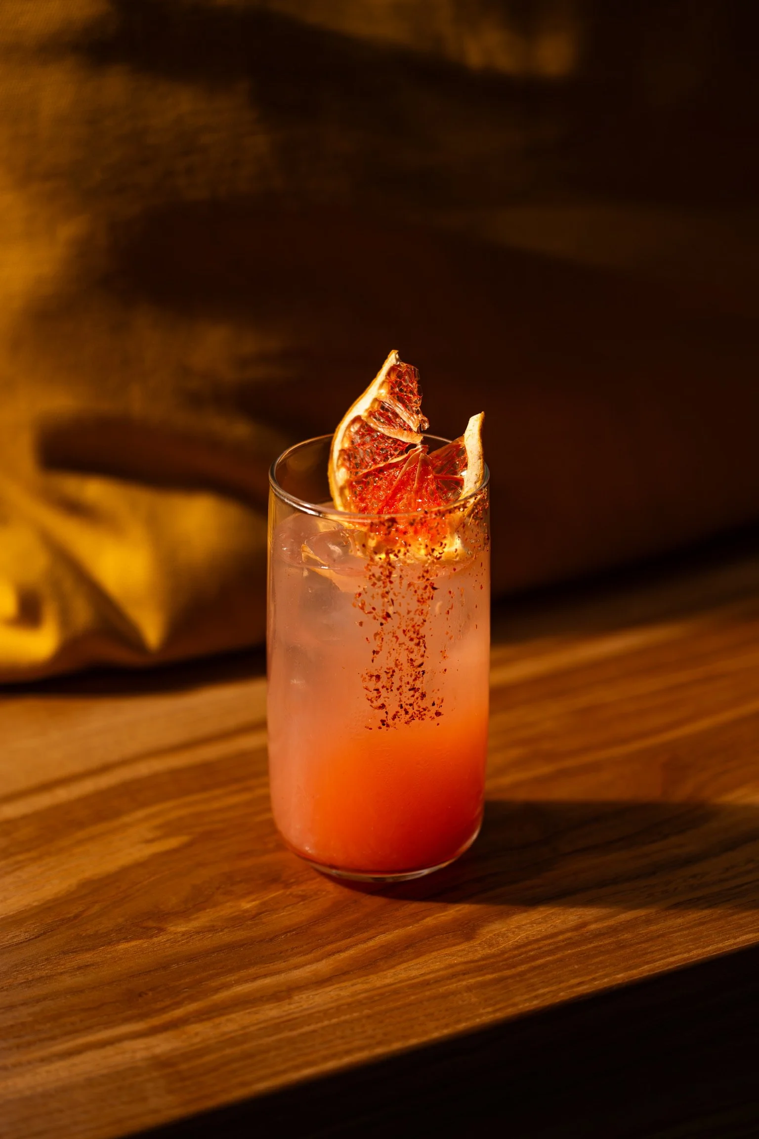 Verre de cocktail rose avec une tranche d'orange séchée et une pincée de poudre rouge en garniture, posé sur une surface en bois.