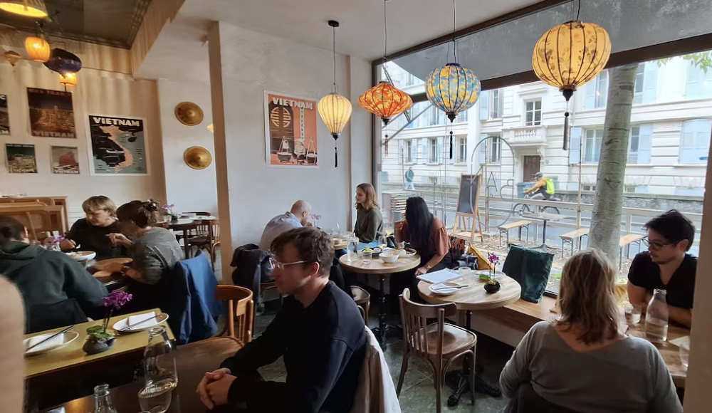 Un restaurant animé avec des clients qui prennent leur repas, décoré avec des lampes colorées et des affiches vintage sur le mur, grande baie vitrée donnant sur la rue