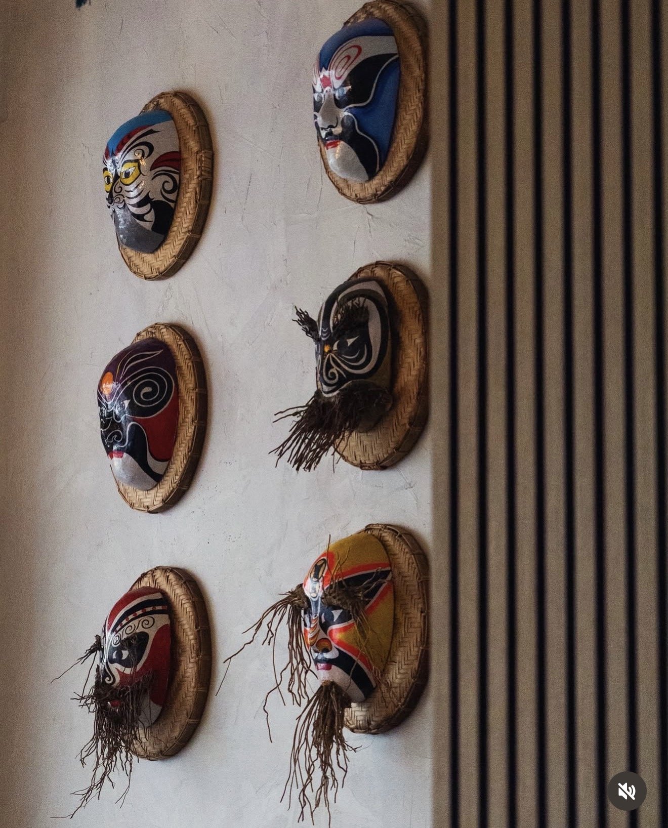 Masques traditionnels asiatiques accrochés à un mur, chacun dans un cadre en rotin, représentant différentes expressions et styles, avec certains ornés de toile de jute et paillettes.
