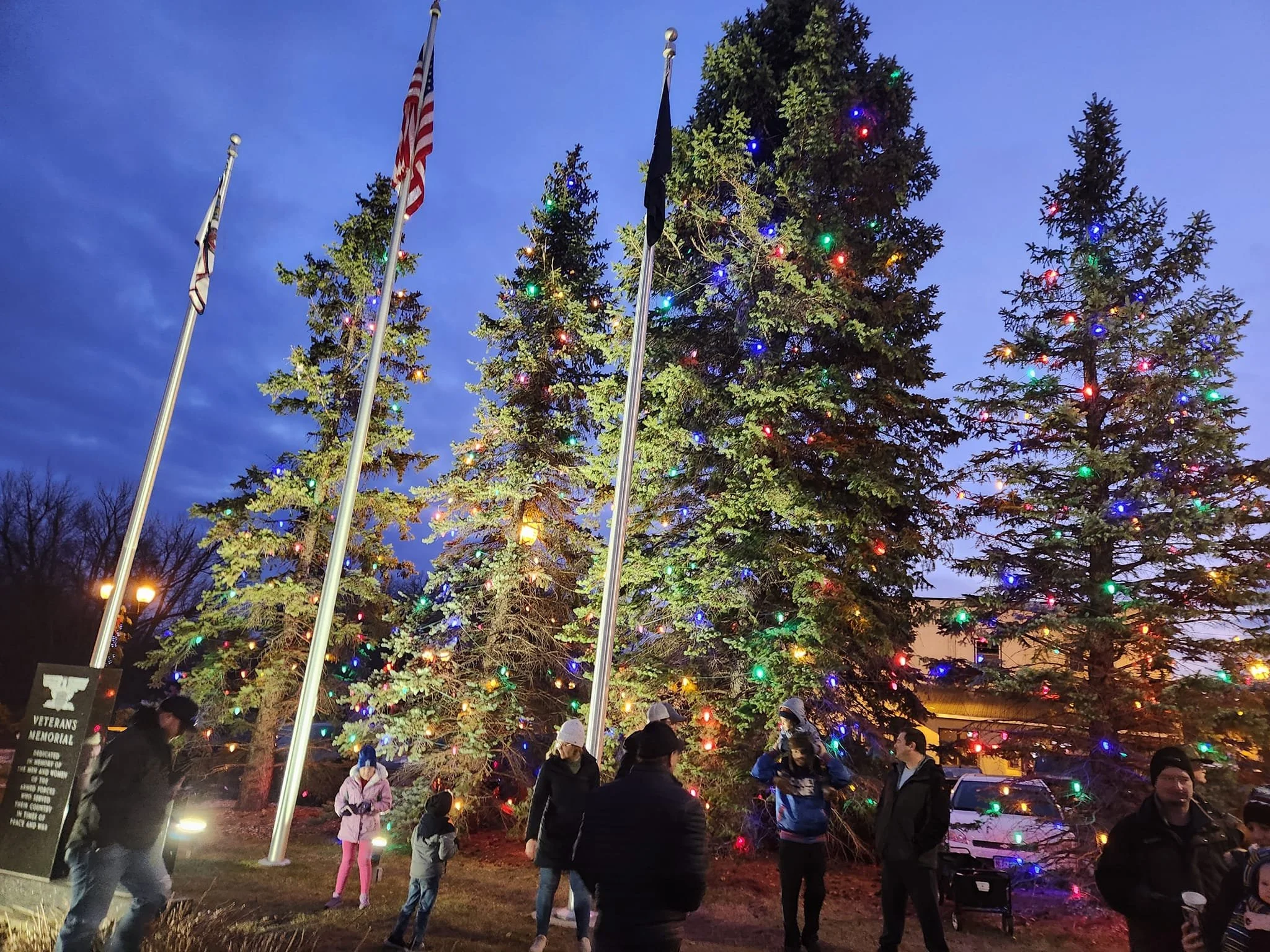 People gathered in front of a large Christmas tree decorated with colorful lights at the Watertown Veterans Memorial during the Christmas tree lighting.