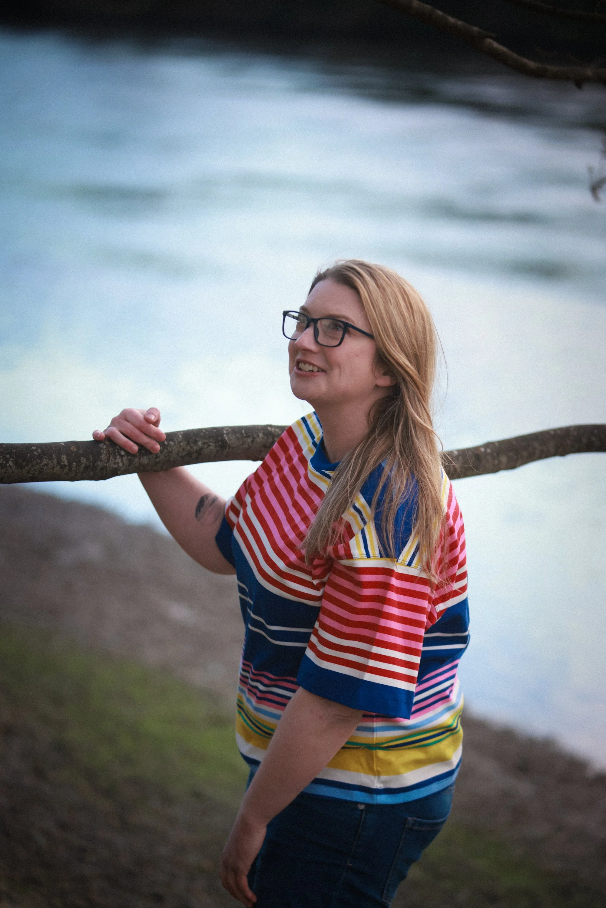 A woman with long hair, glasses, and tattoos on her arm, wearing a multicolored striped shirt, stands outdoors near water, holding onto a tree branch and smiling.