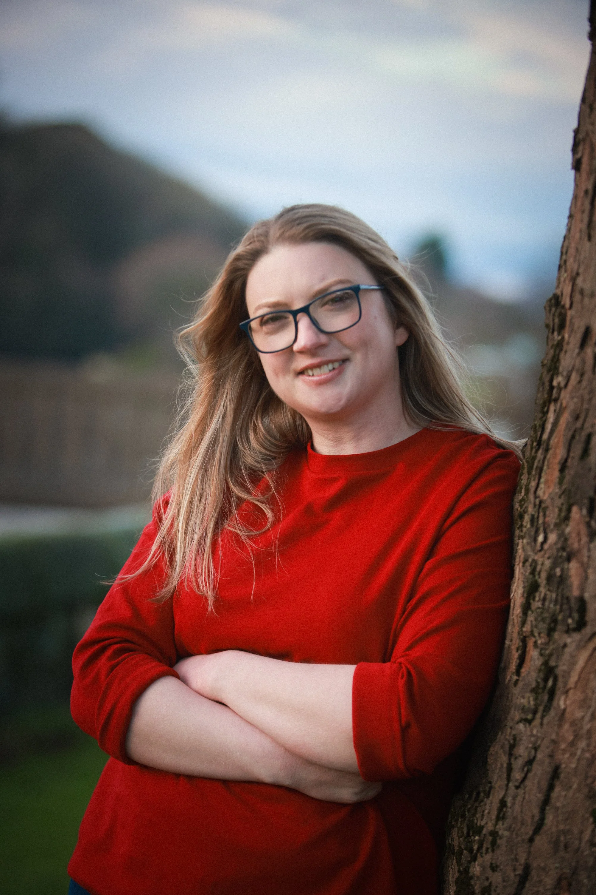 A woman with long blonde hair, wearing glasses and a red long-sleeve shirt, leaning against a tree and smiling outdoors during the daytime.