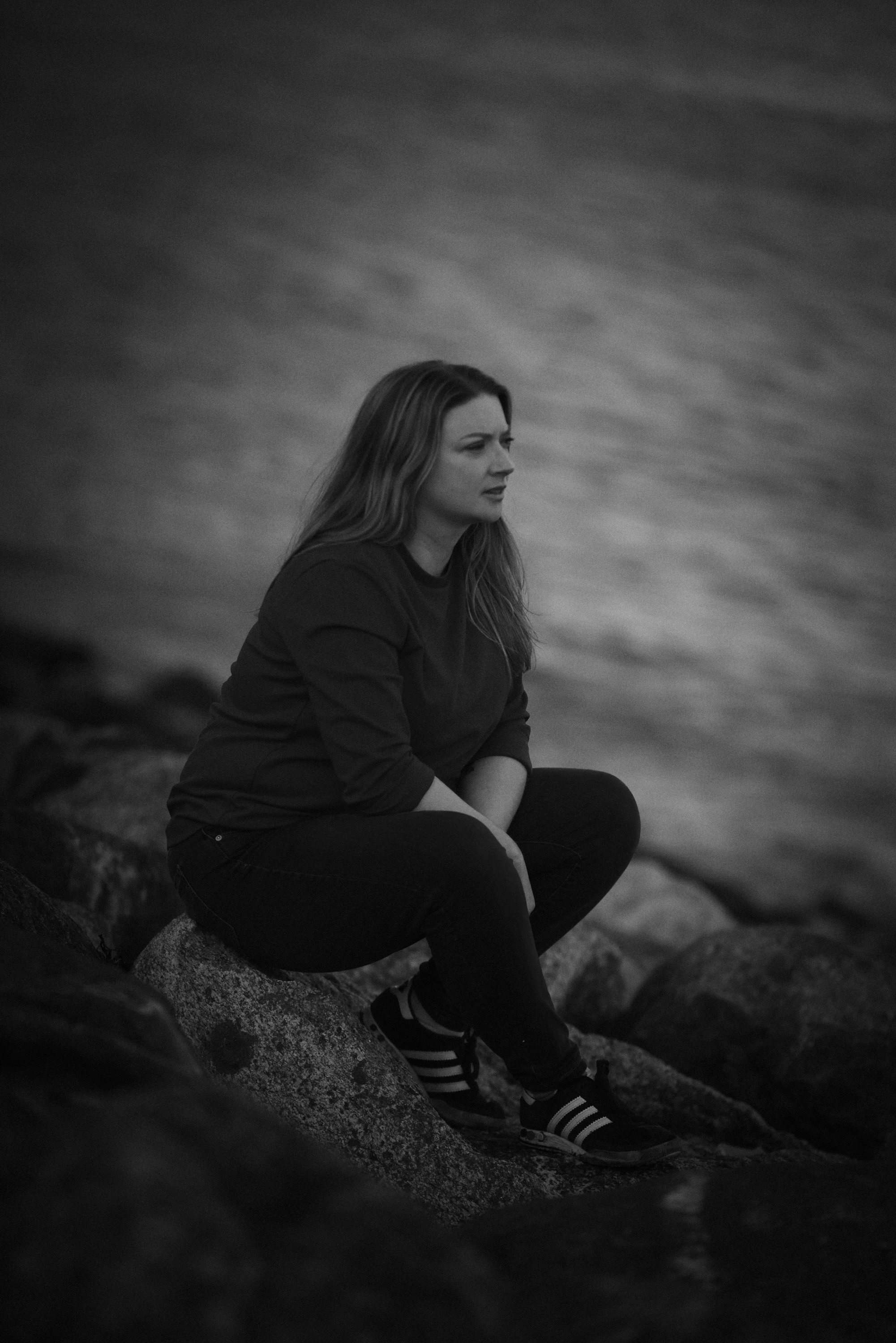 A woman sitting on rocks near water, looking thoughtful or concerned, in black and white.