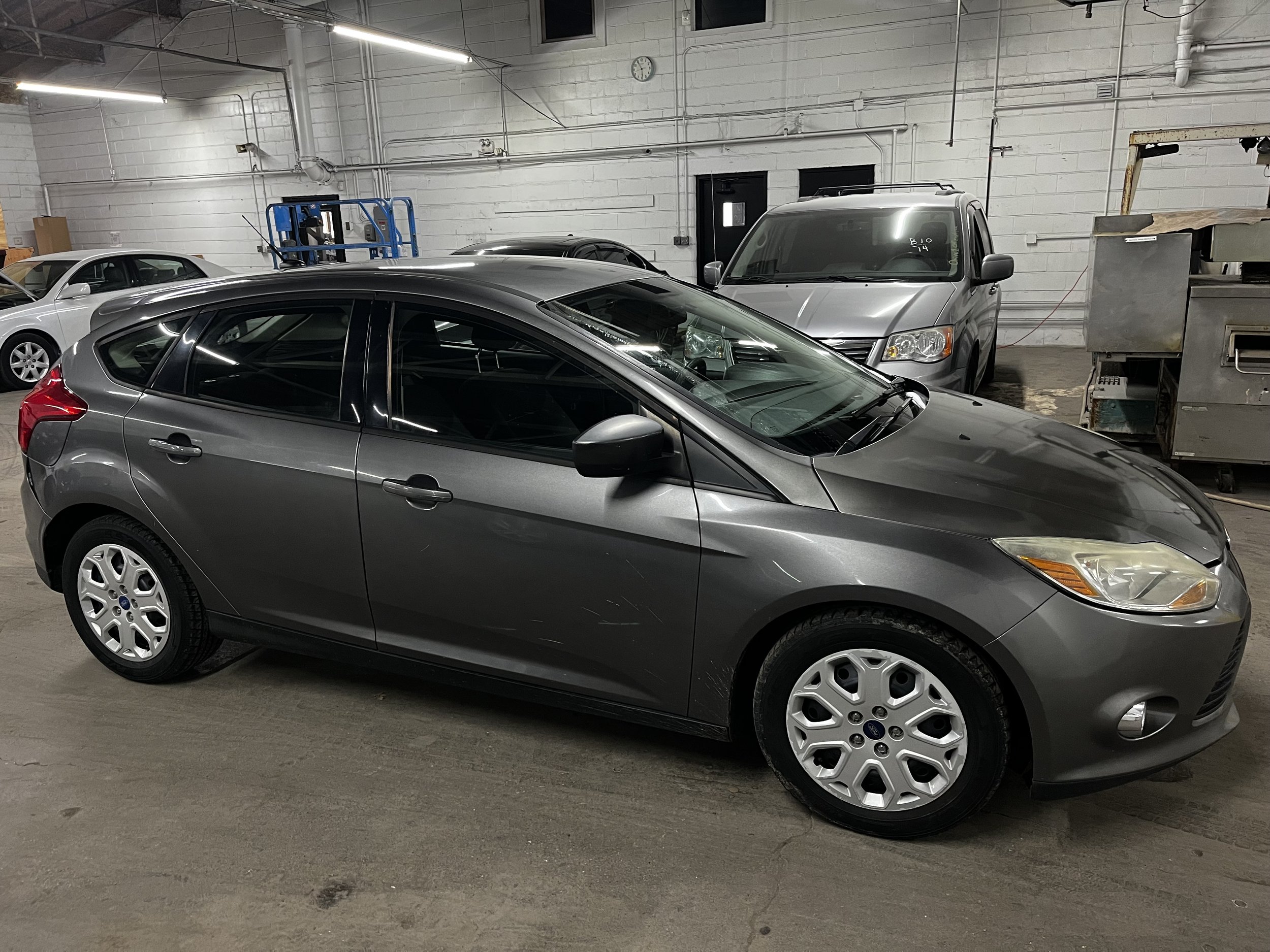 Gray hatchback car parked in a garage with other vehicles in the background.