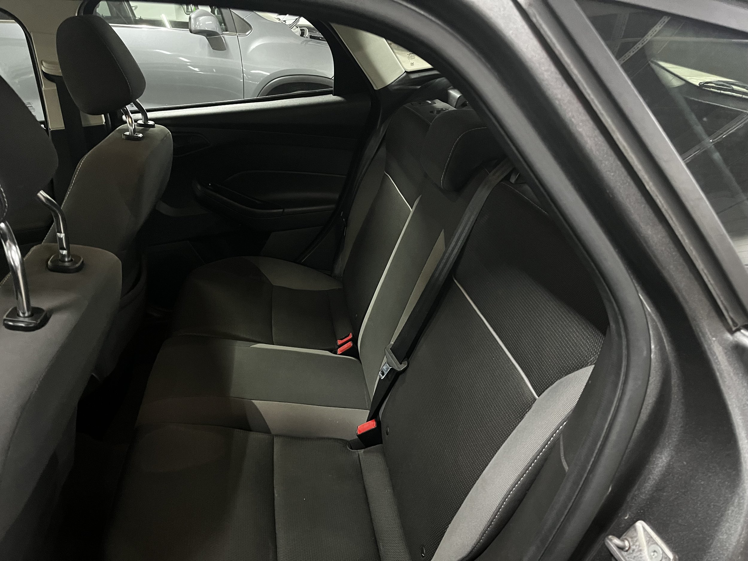 Interior of a car showing the back seats with black upholstery and red seatbelt buckle clips. The front seats are partially visible with adjustable headrests. The car door is open, revealing the side of a silver vehicle parked nearby.