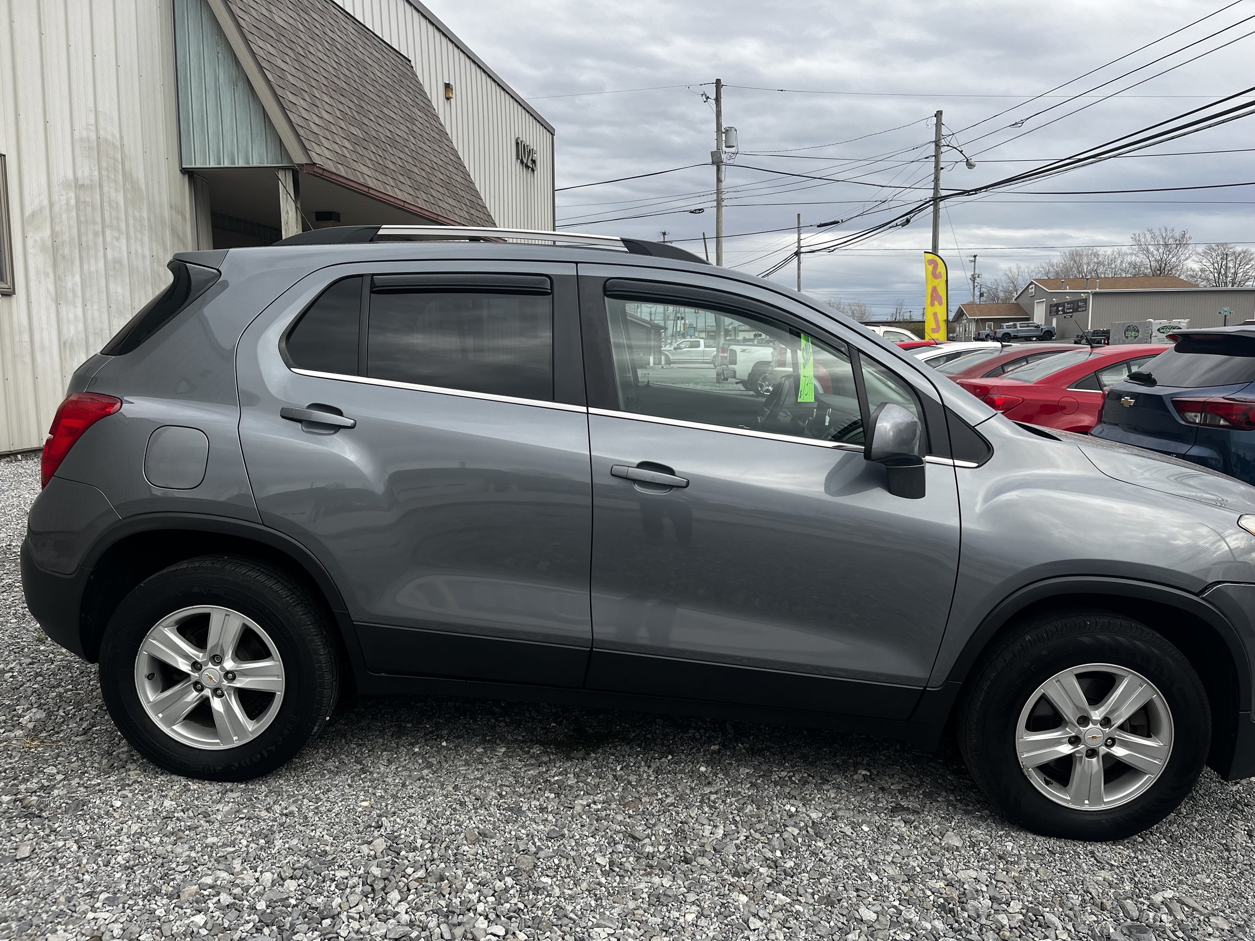 Gray SUV parked on gravel near a building with a sloped roof, surrounded by other cars on a cloudy day.