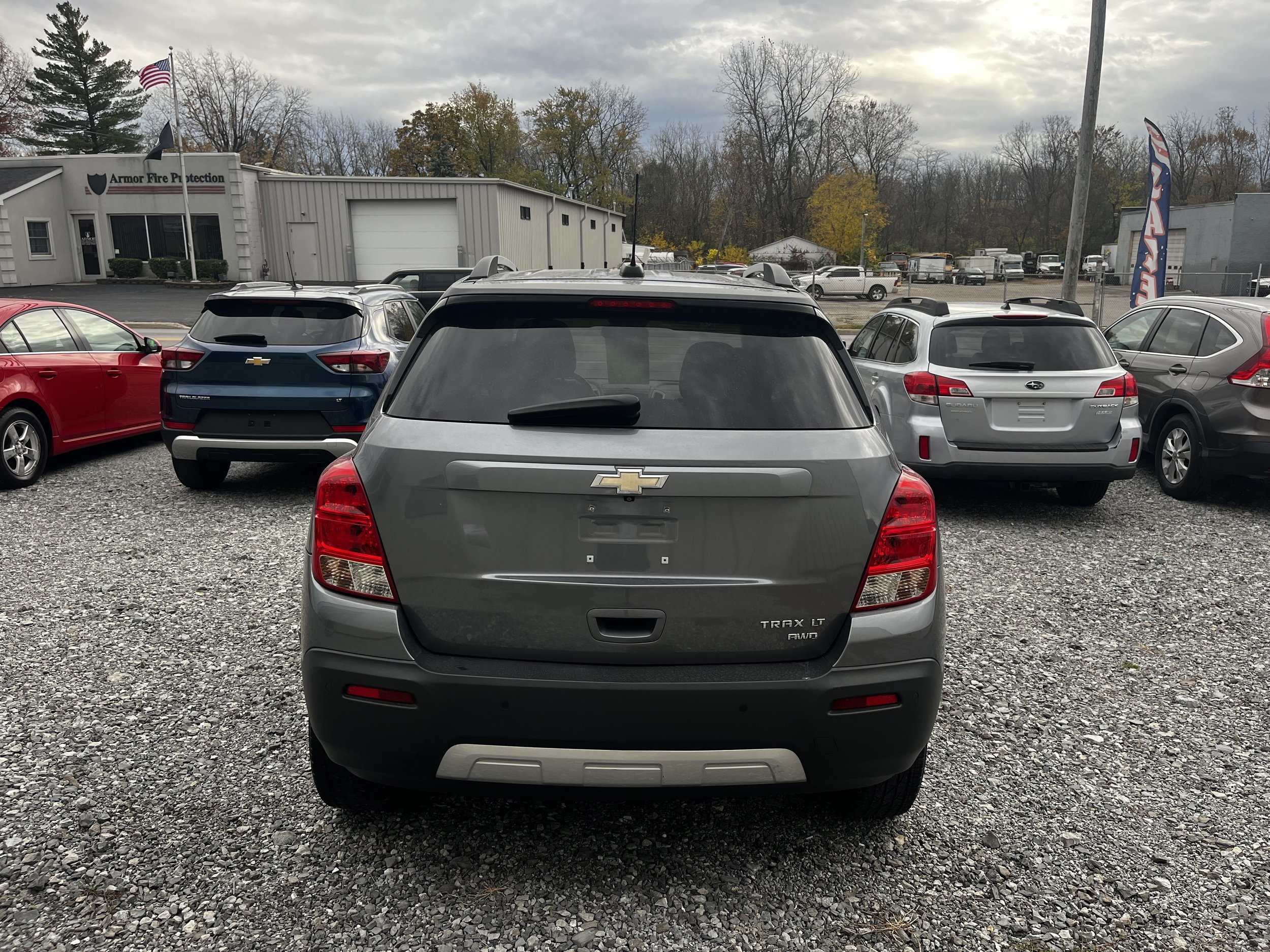 Back of a gray Chevrolet Trax LT parked among other cars on a gravel lot, near a building with an American flag visible.