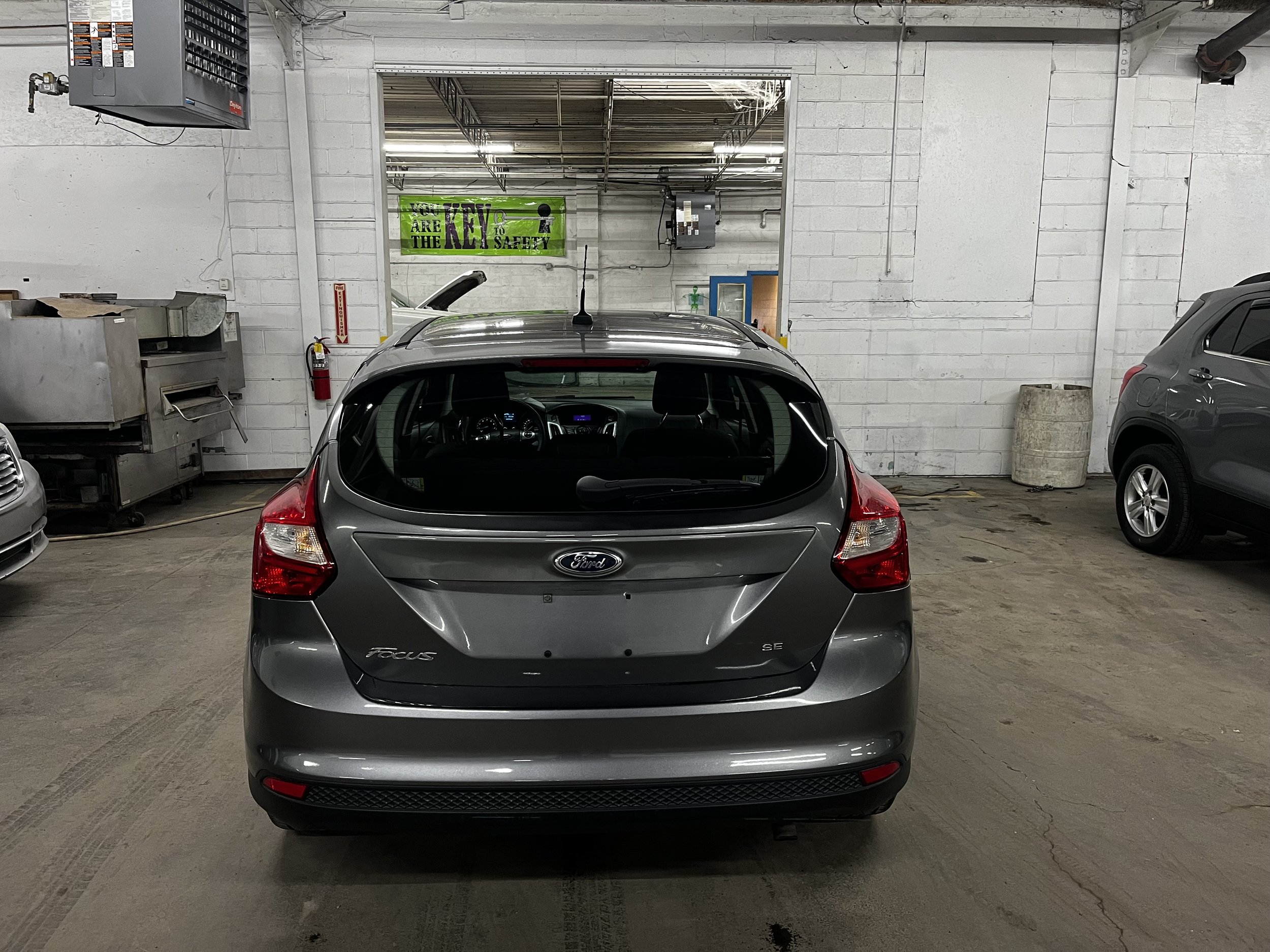 Rear view of a gray Ford Focus SE car in a garage with other vehicles and industrial equipment.