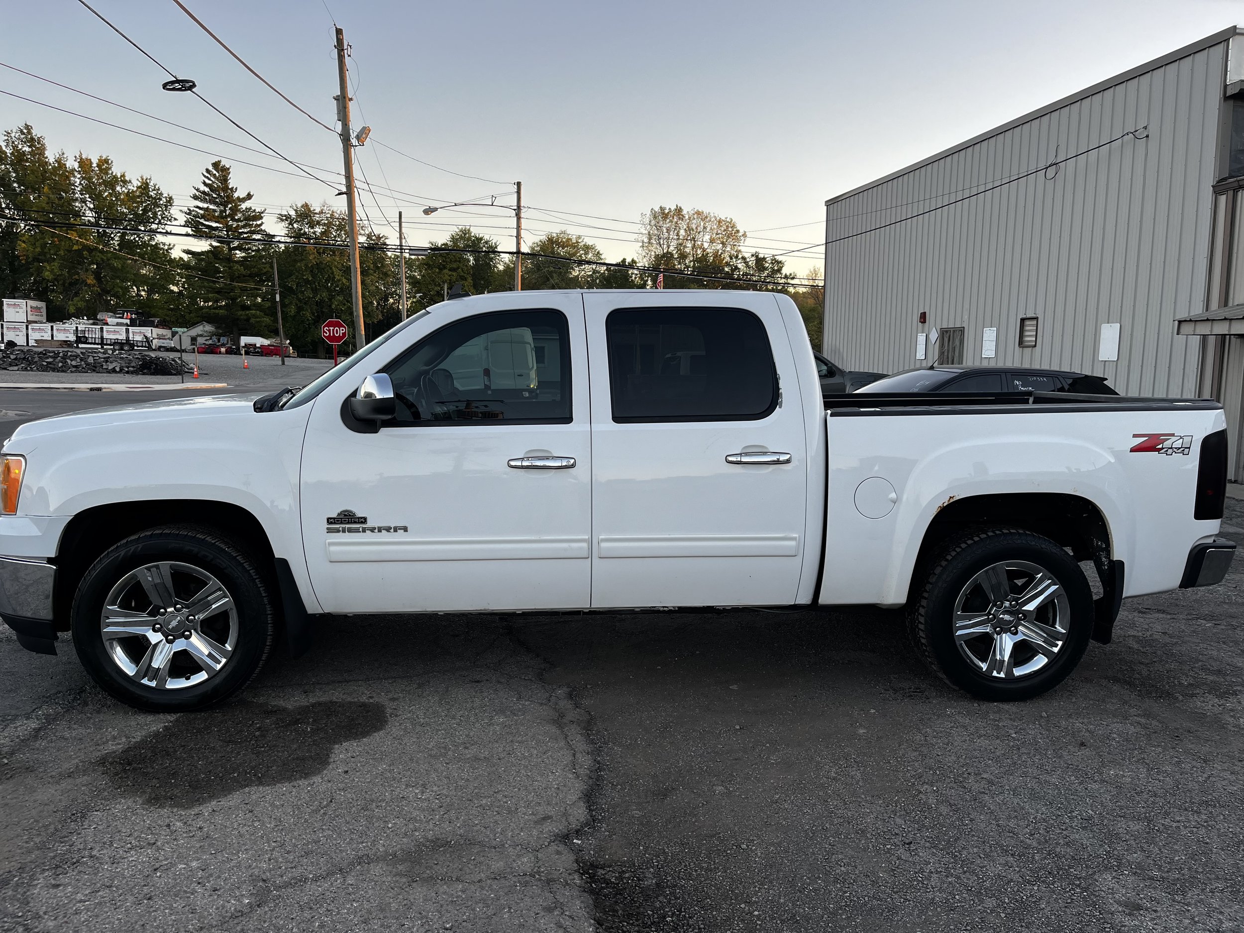 White GMC Sierra pickup truck parked outdoors in front of an industrial building, with trees and power lines in the background.