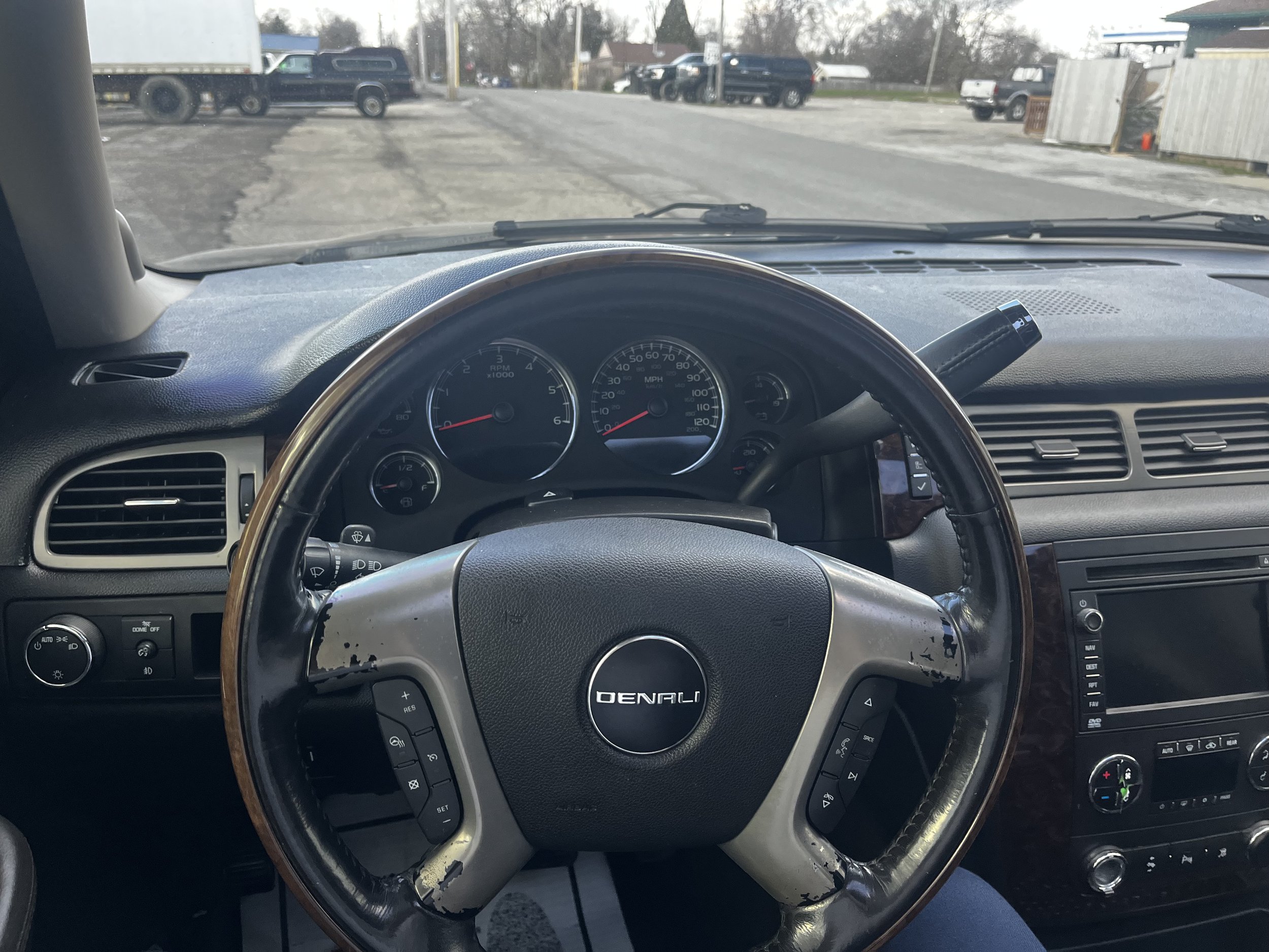 Interior view of a vehicle dashboard showing a Denali steering wheel, speedometer, and other control gauges. There's also a view through the windshield showing a parking lot with several trucks. The dashboard includes a radio and air vents.