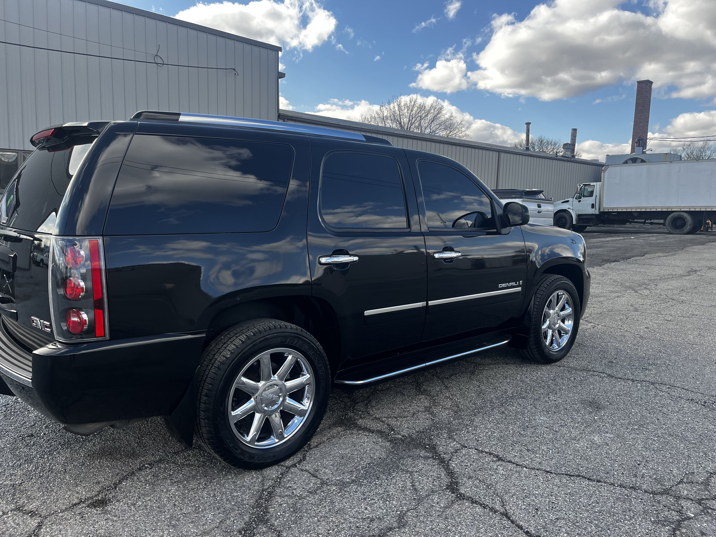 Black GMC Denali SUV parked in an industrial area with other vehicles and a building in the background.